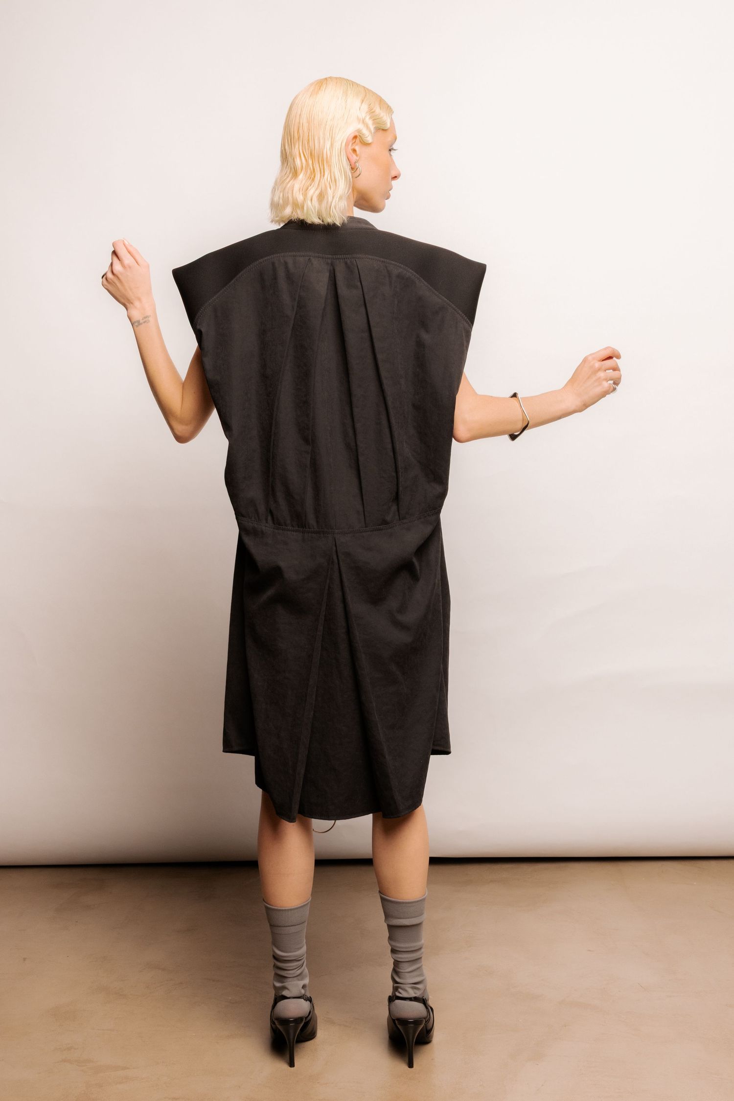 Back view of a black oversized sleeveless dress with structured shoulders and pleated detail, worn with grey socks and black heels.