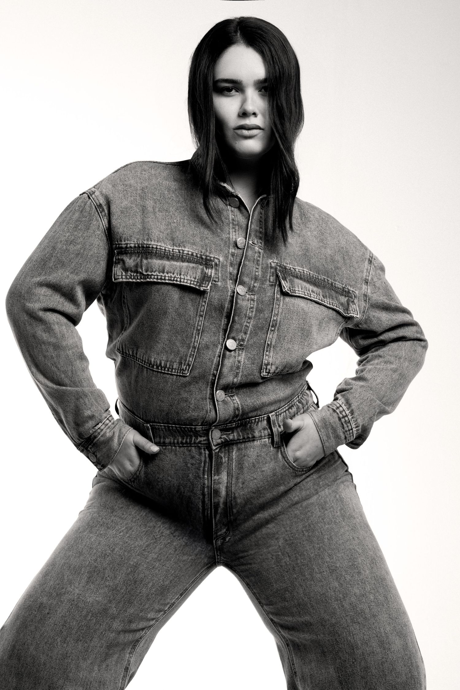 Black and white image of a model in a denim jumpsuit standing with hands on hips and a confident, direct stare.