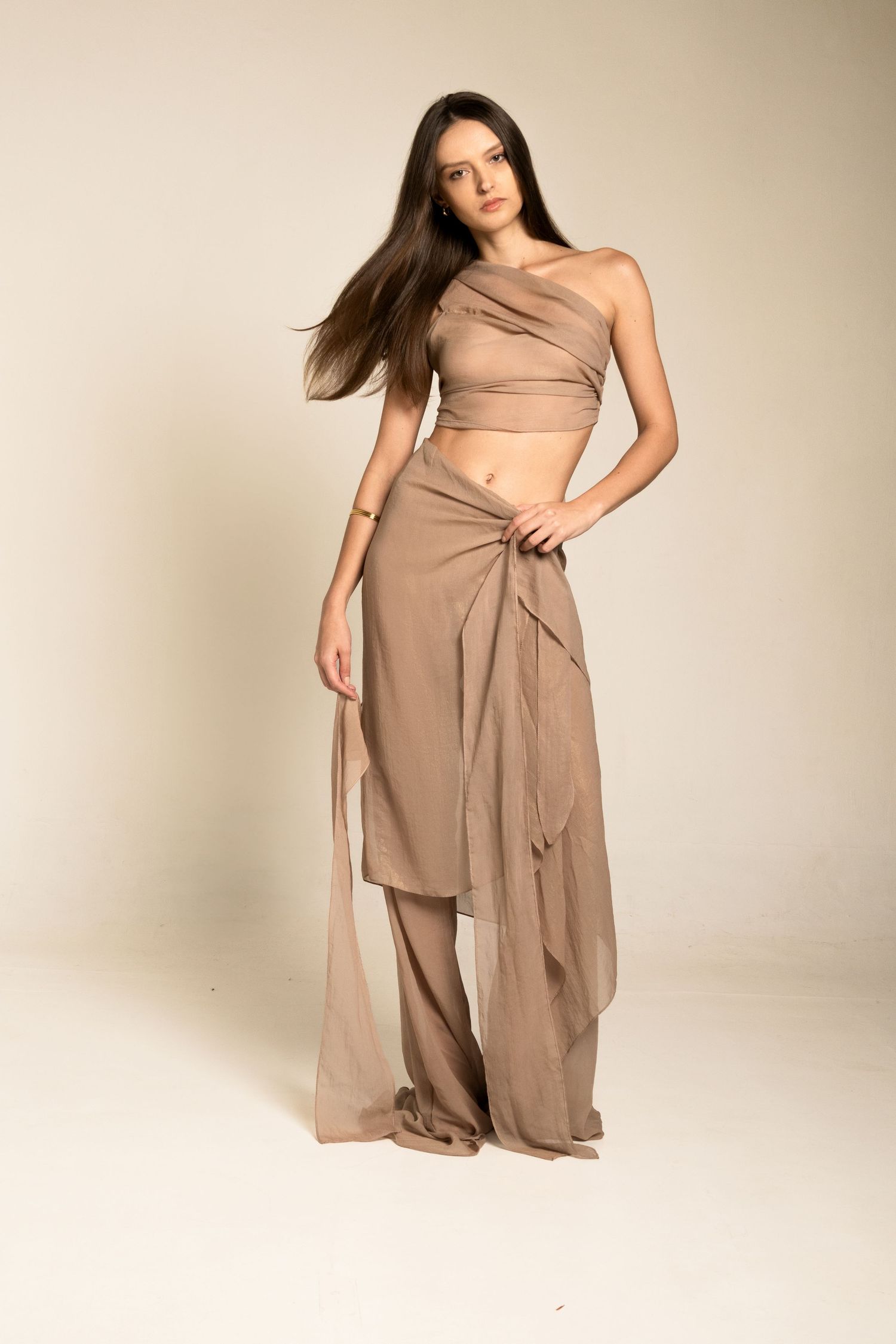 Full-length studio shot of a model wearing a taupe asymmetric crop top and draped sheer wrap skirt over wide-leg pants.