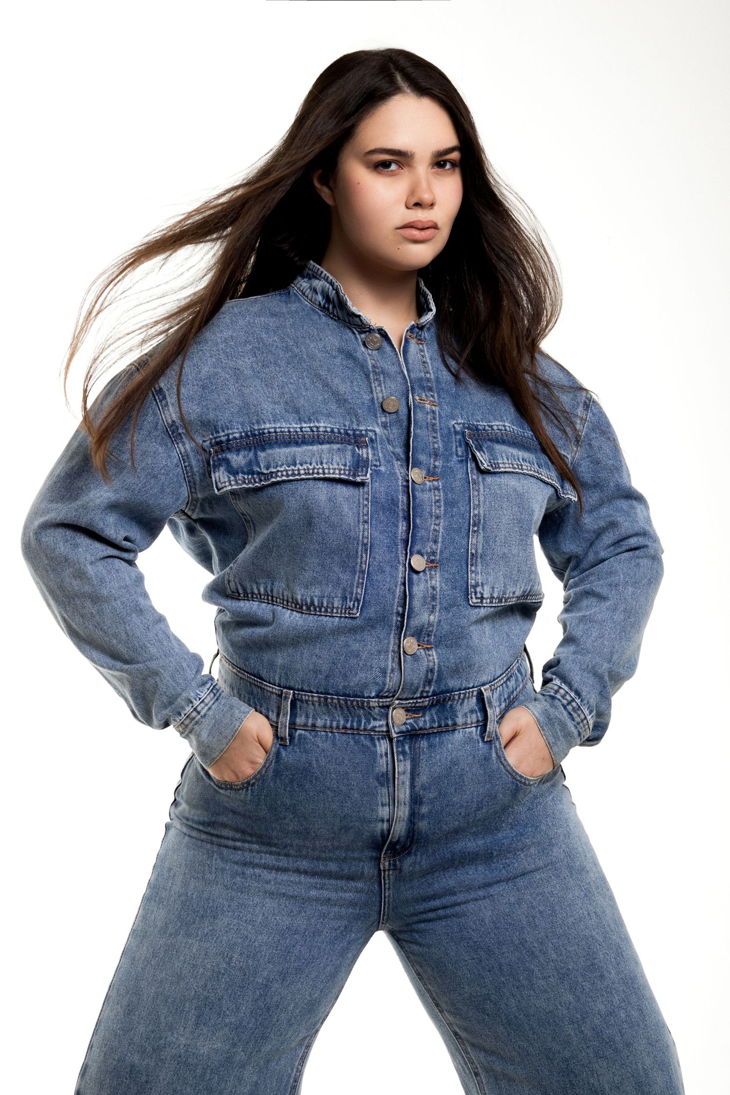 Model with long flowing dark hair wears a blue denim jumpsuit with hands in pockets and gazes intensely at the camera.