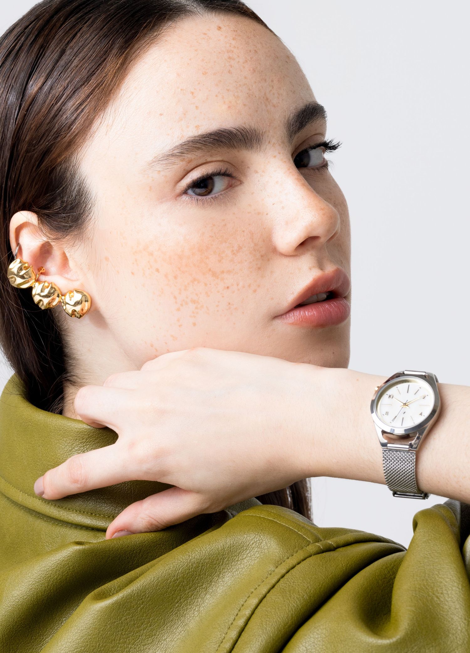 Woman with a gold ear cuff and olive leather jacket rests her hand at her neck, wearing a silver mesh strap watch with a white dial.