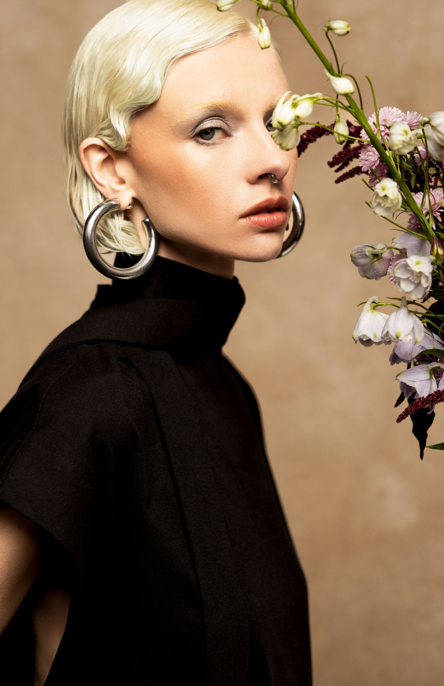 Close-up portrait of a platinum blonde model in a black high-neck top and chunky silver hoop earrings beside delicate pink flowers.