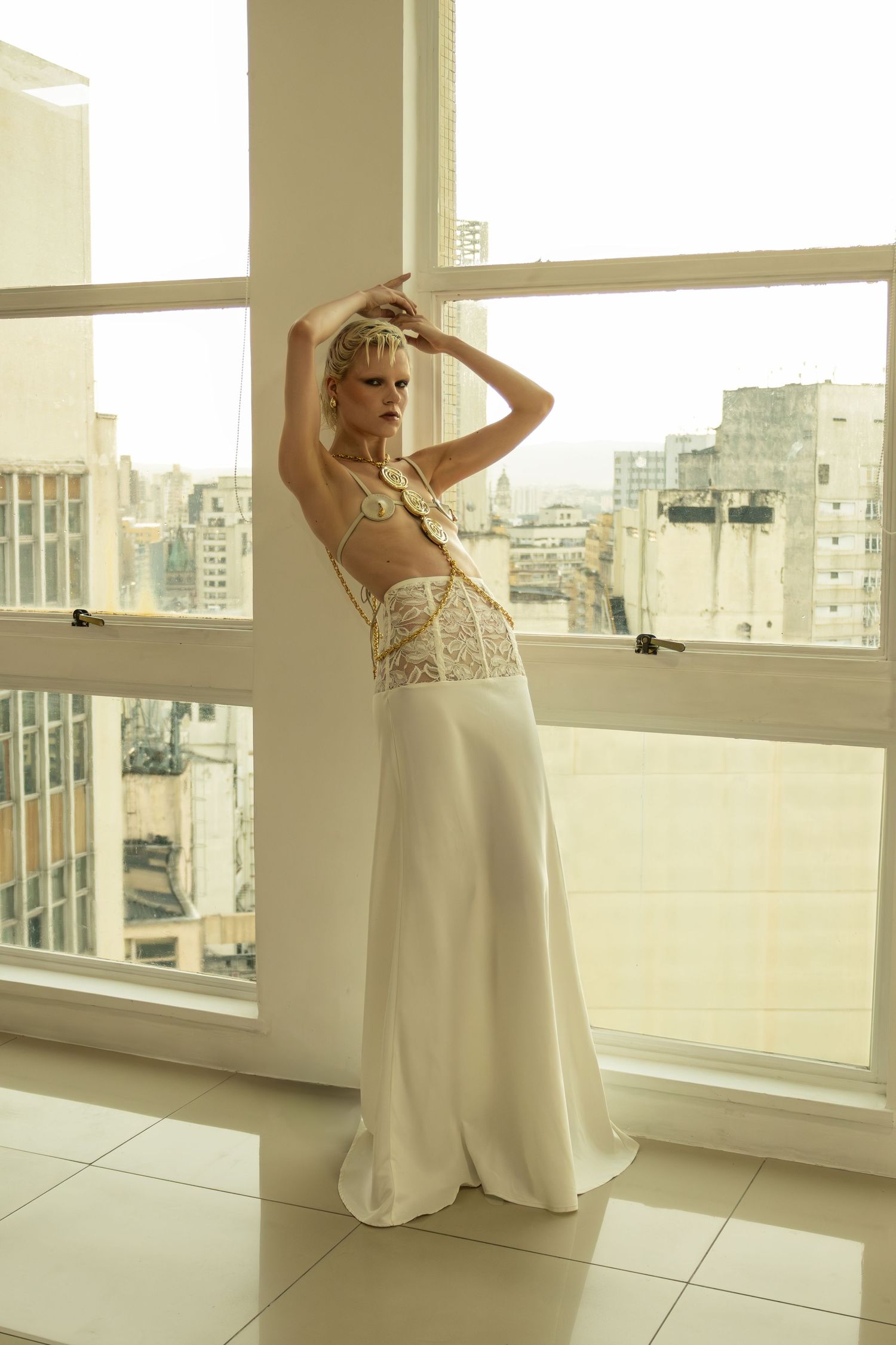 A model in a long white skirt and gold chain top poses with arms raised beside large windows overlooking a city skyline.