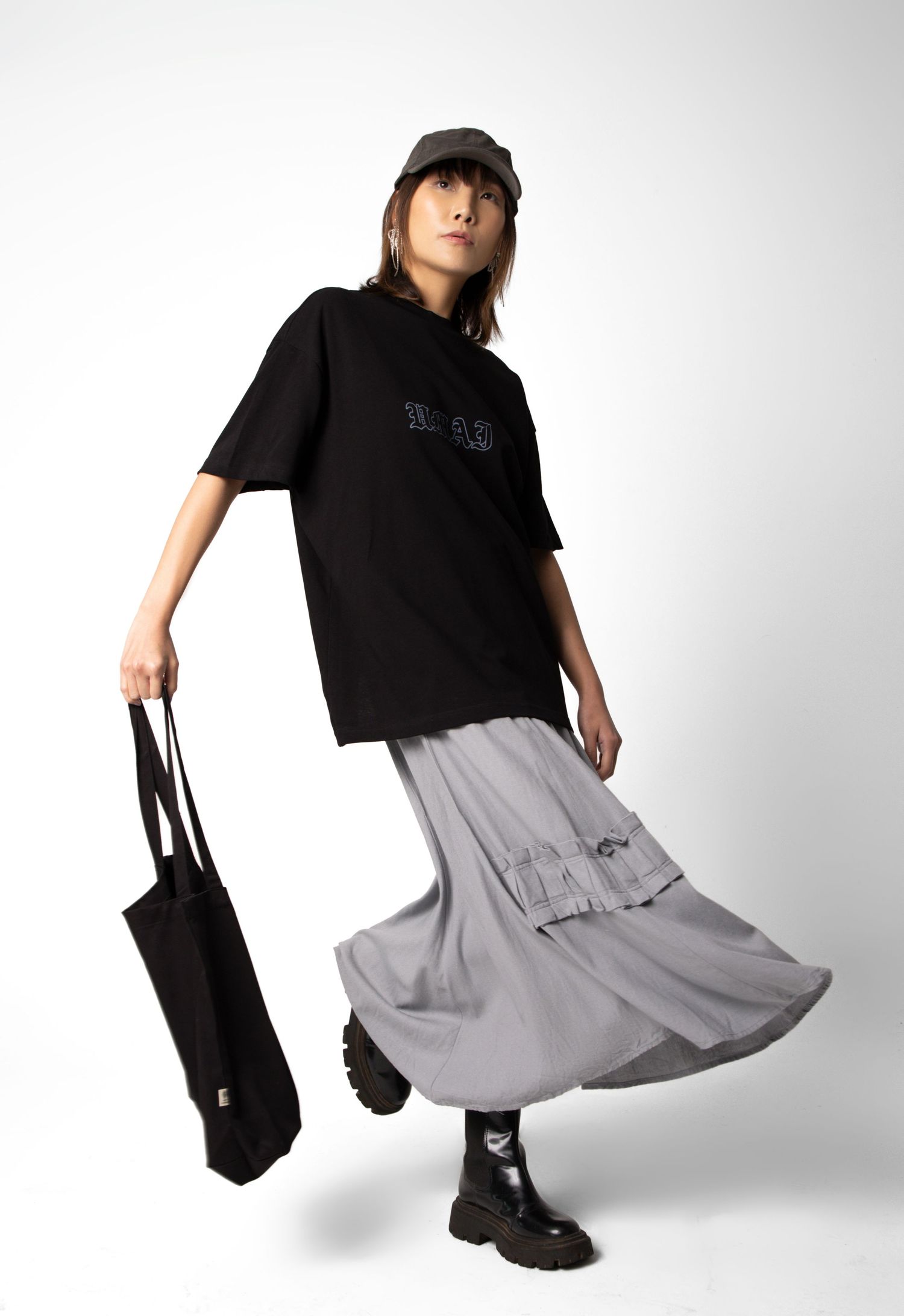 Woman in a black oversized tee, gray cargo maxi skirt, cap and chunky black boots walks while holding a black canvas tote bag.