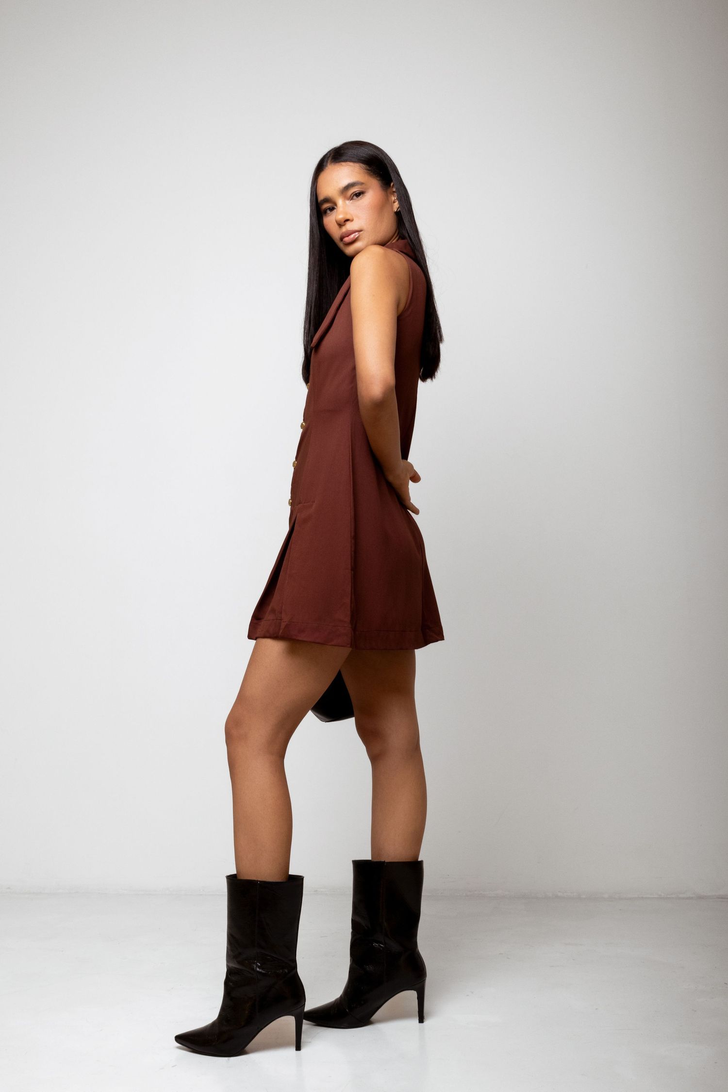 Side view of a woman in a brown sleeveless mini dress with a flared hem and black pointed heeled ankle boots against a white backdrop.