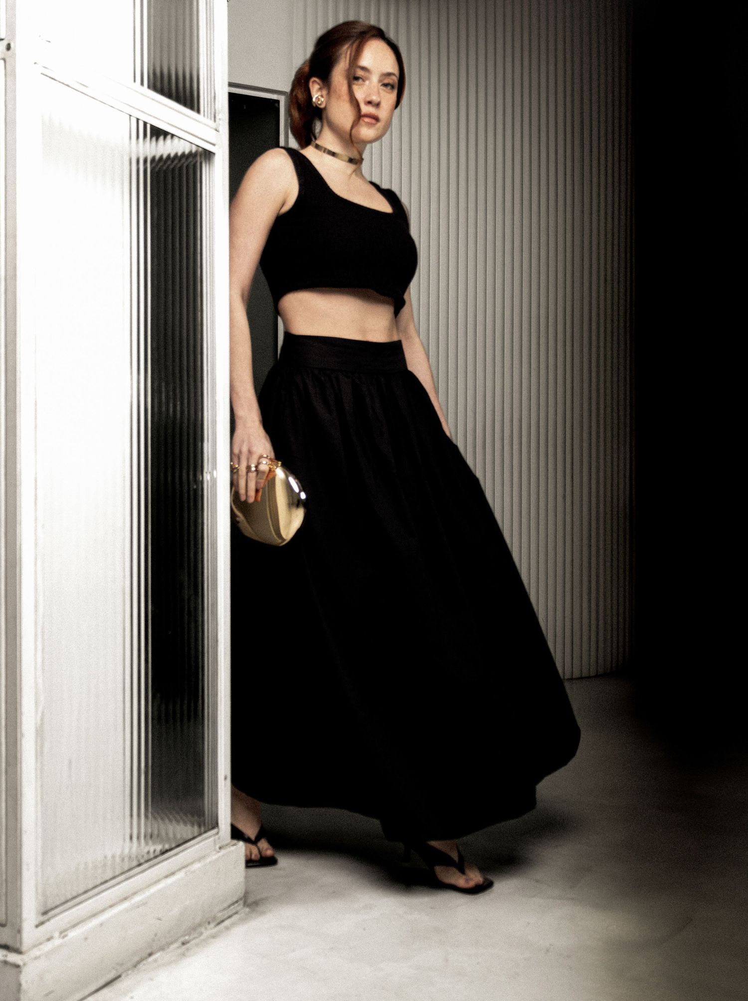 Model poses by a ribbed glass door in a black crop top, flowing maxi skirt, and sandals, carrying a gold clutch.