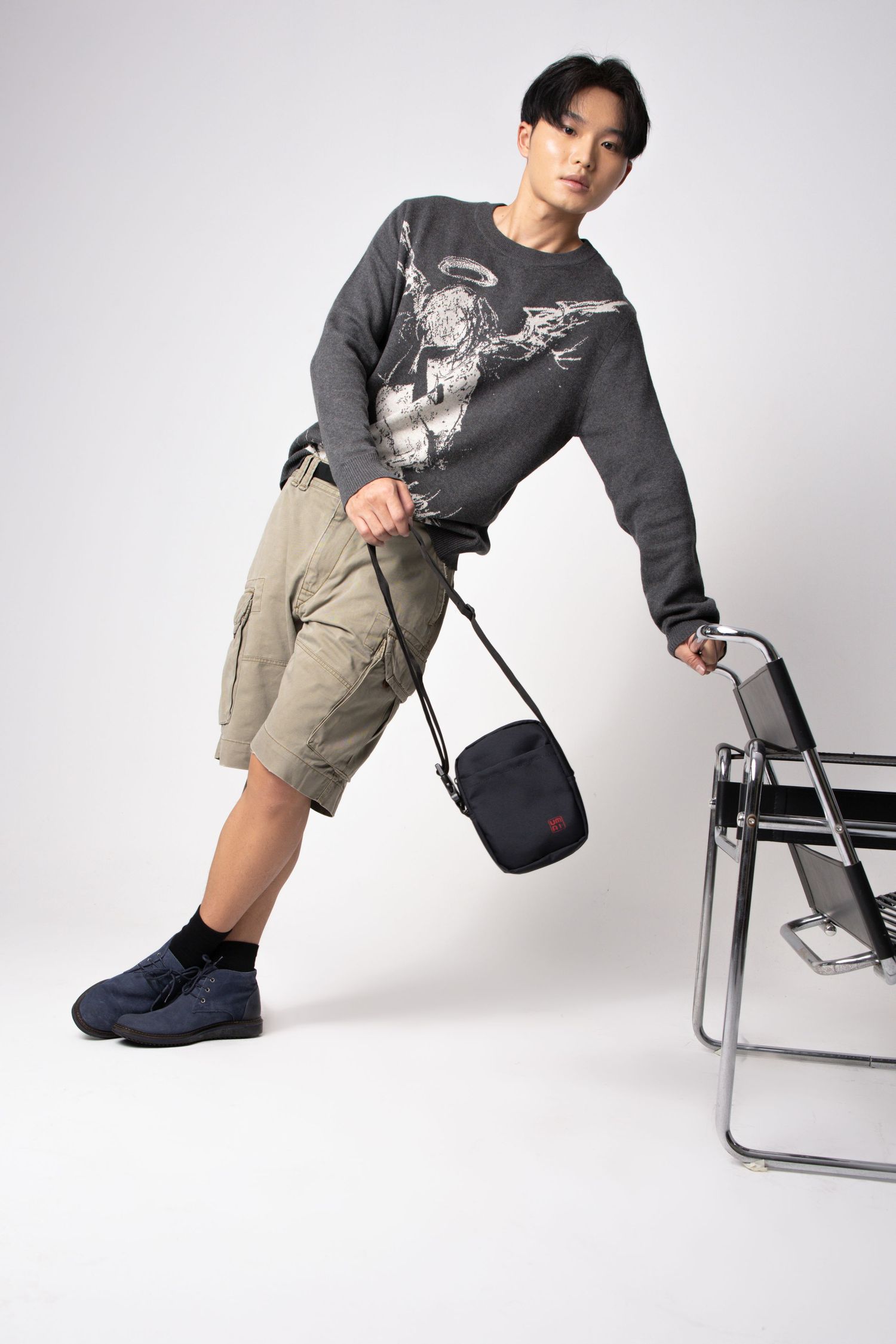 Young man in a graphic sweater and cargo shorts leans on a chrome chair holding a small black crossbody bag.