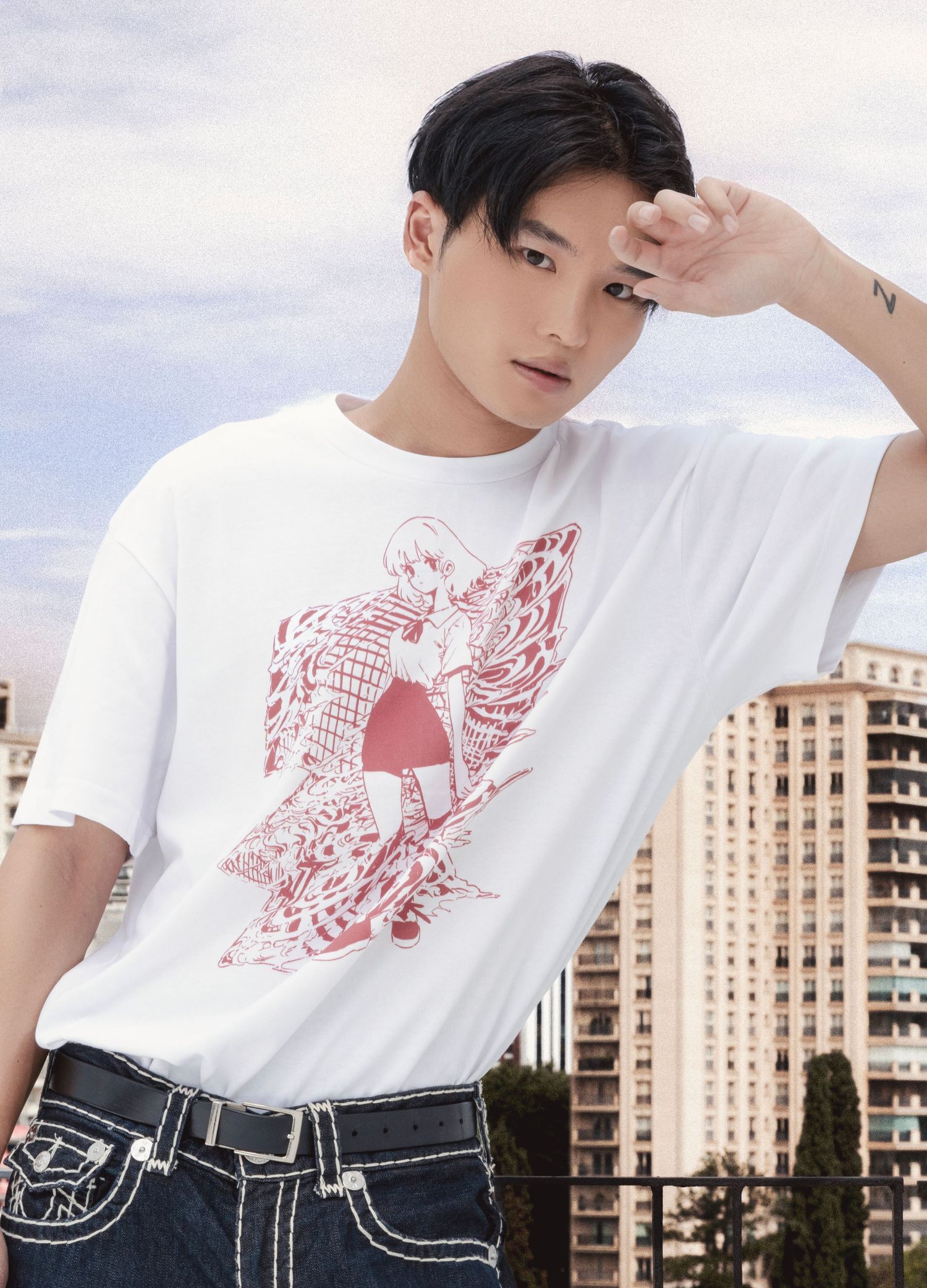 Young man with dark hair raises his arm wearing a white graphic anime-print t-shirt and jeans in front of city apartment buildings.