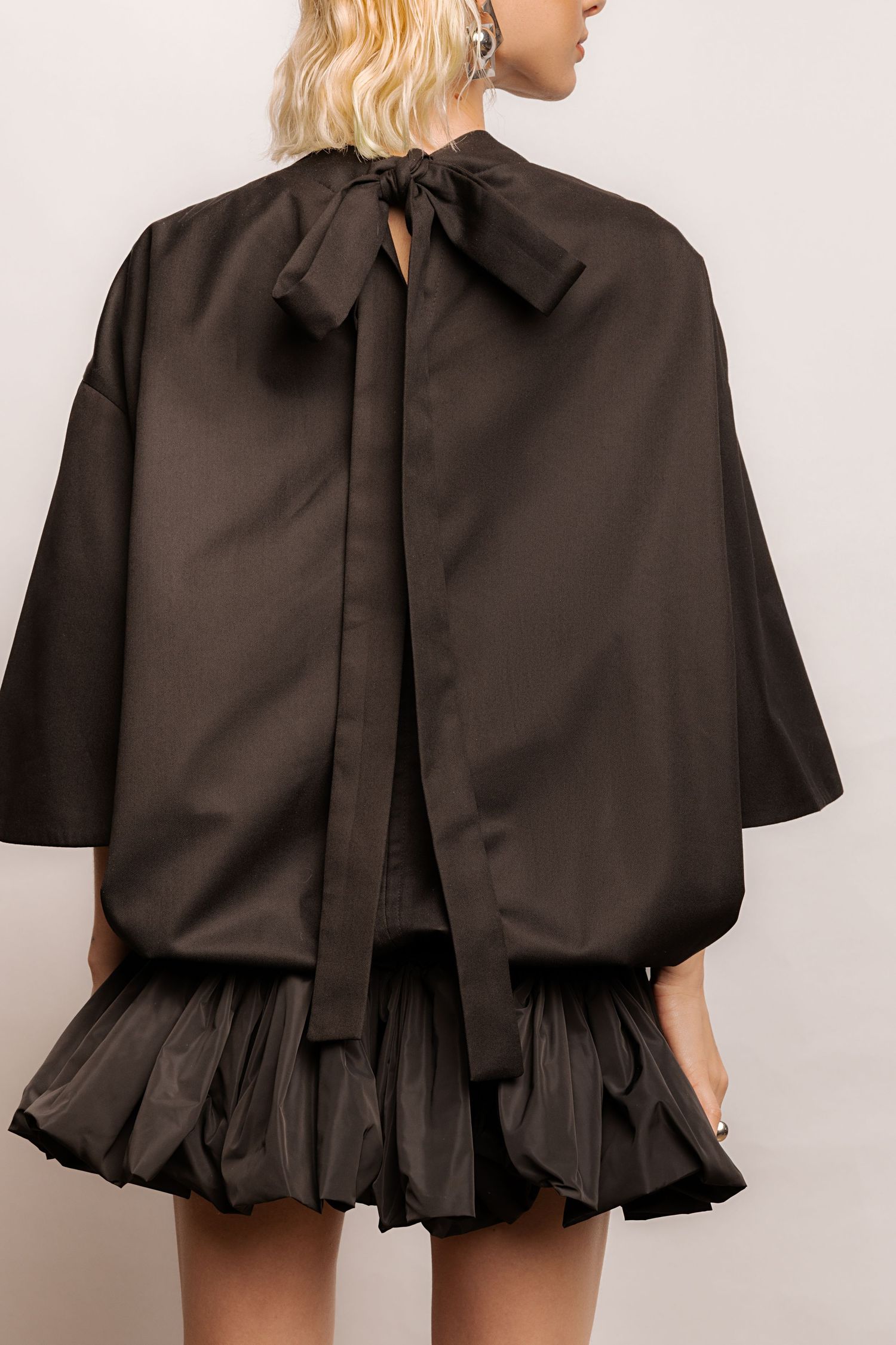 Back view of a model in a black oversized top with a long tie bow at the neck and a black ruffled bubble-hem mini skirt.