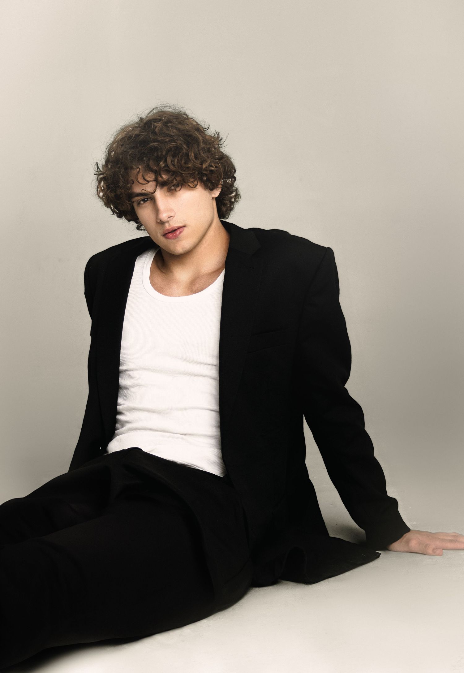 Curly-haired model sits on the studio floor in a black oversized blazer and white tank top, gazing at the camera.