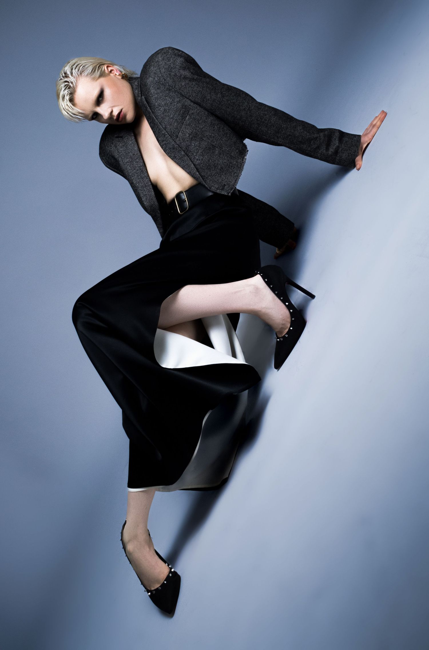 Model in a gray tweed jacket, black wide-leg trousers and studded black heels strikes a dynamic angled pose against a blue-gray backdrop.