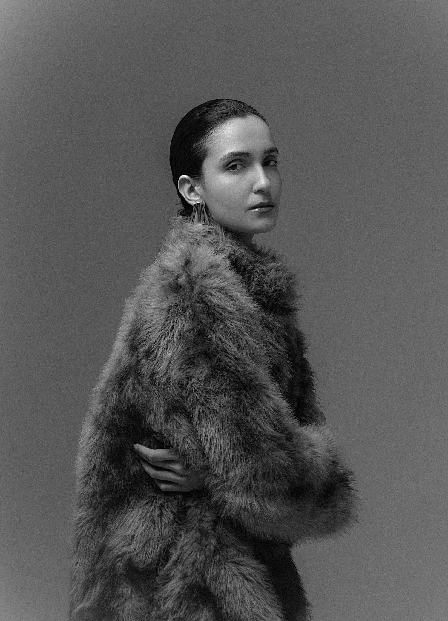 Black and white portrait of a model with slicked-back hair wearing a plush faux fur coat and drop earrings.
