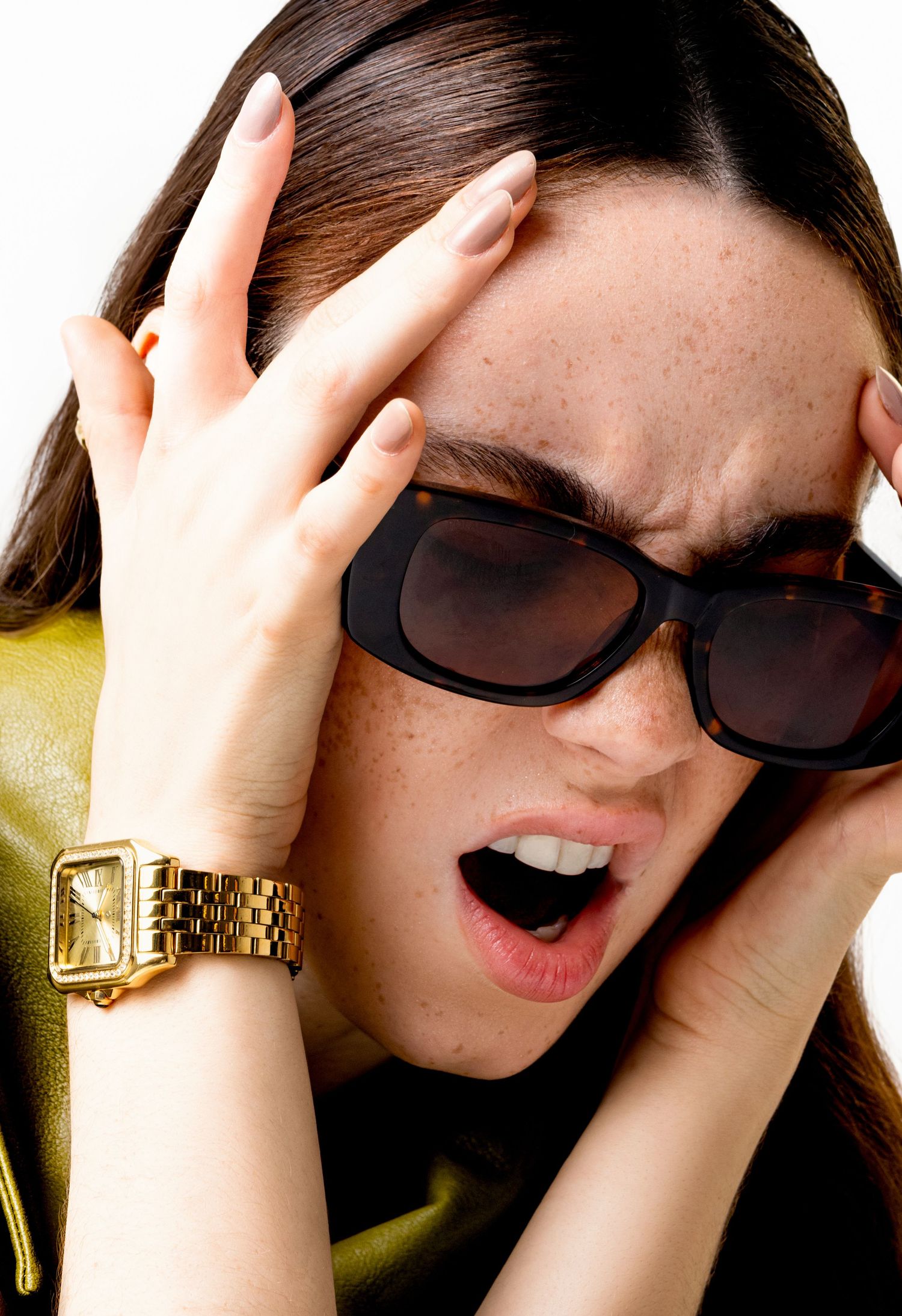 Woman in dark sunglasses grips her head with an expressive look, wearing a gold rectangular bracelet watch and an olive jacket.
