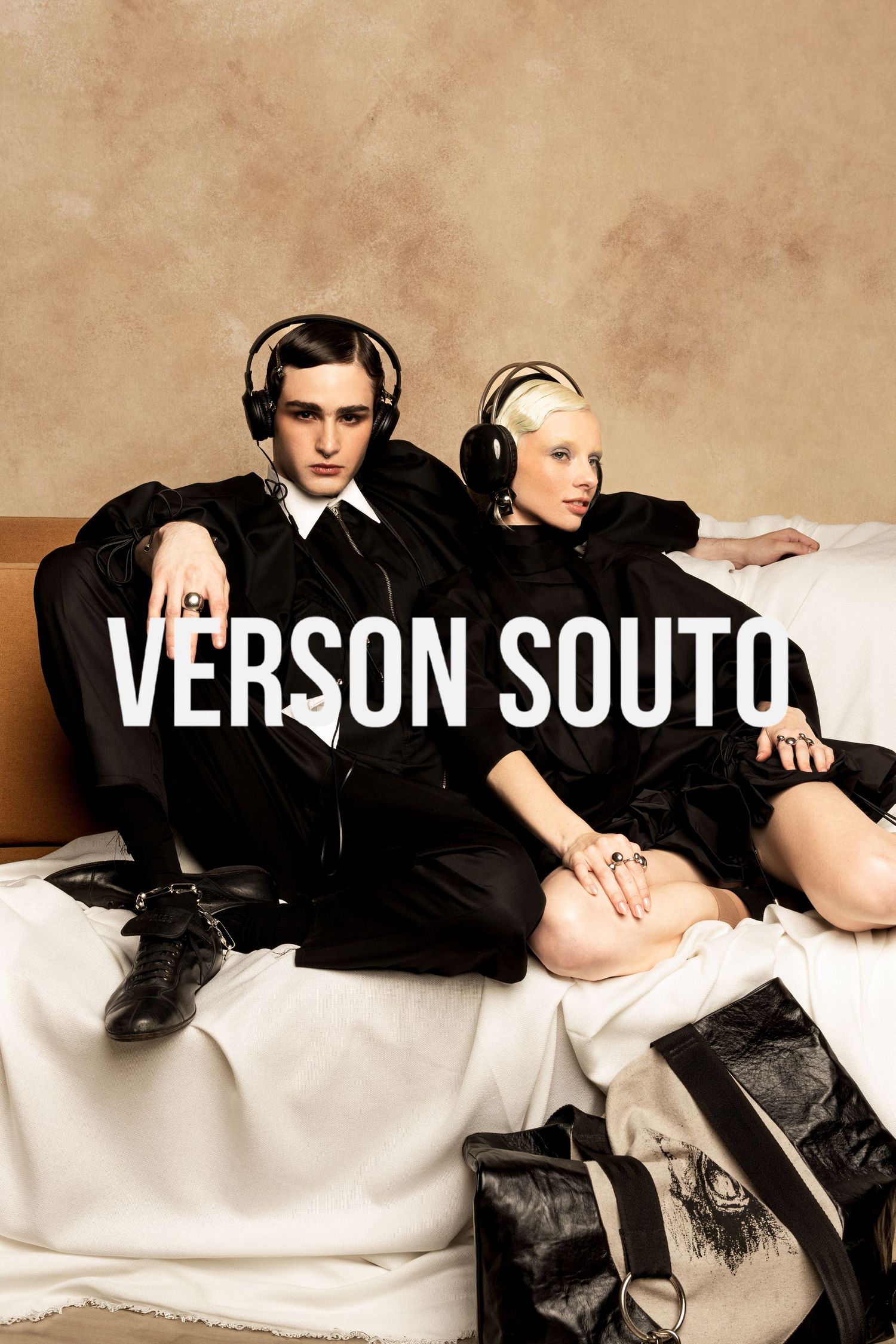 Verson Souto fashion campaign showing two models in black outfits and headphones on a sofa with a black leather bag in the foreground.