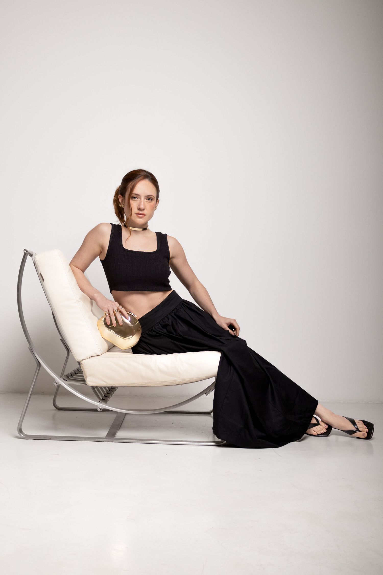 Woman in a black crop top and long black skirt sits on a sleek cream and metal chair holding a gold clutch in a white studio.