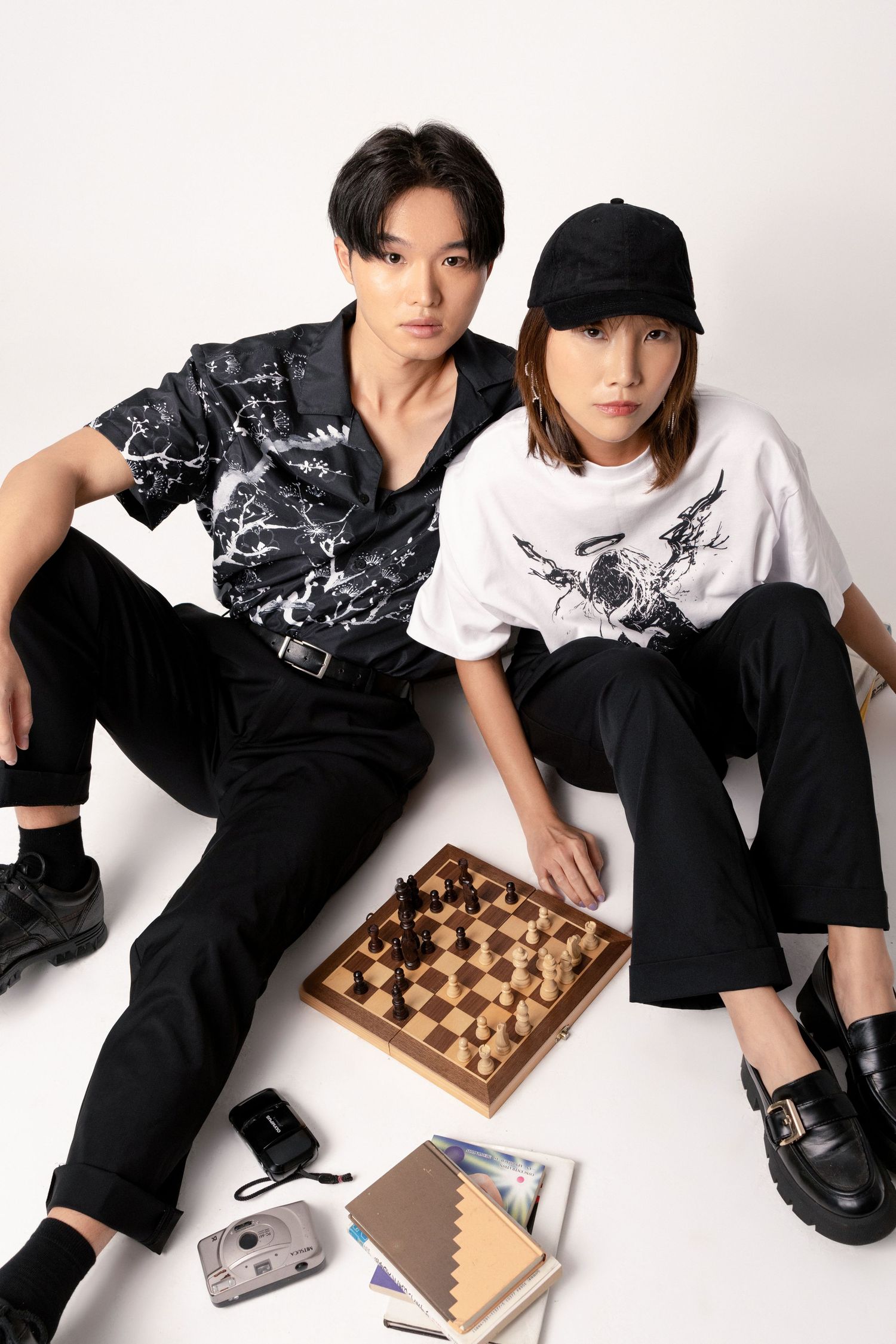 Two models in black and white streetwear sit on a white floor beside a wooden chess set, a film camera and books.