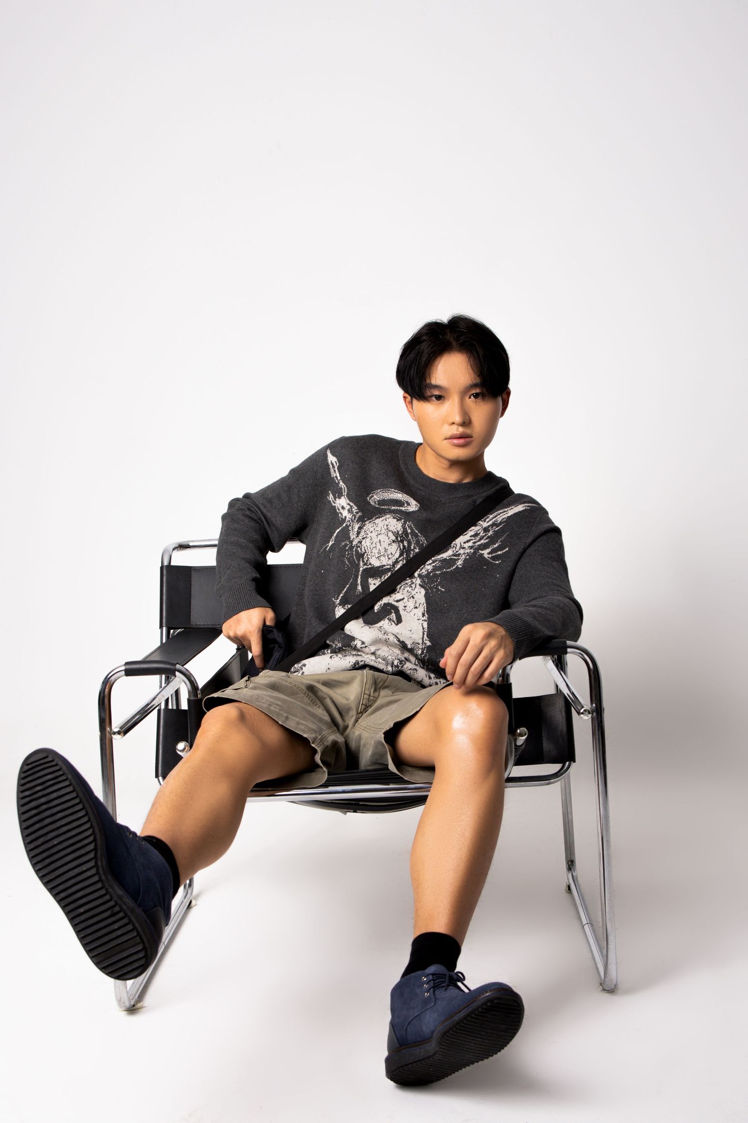 Young man in a graphic sweater and cargo shorts sits in a chrome and leather chair against a white backdrop.