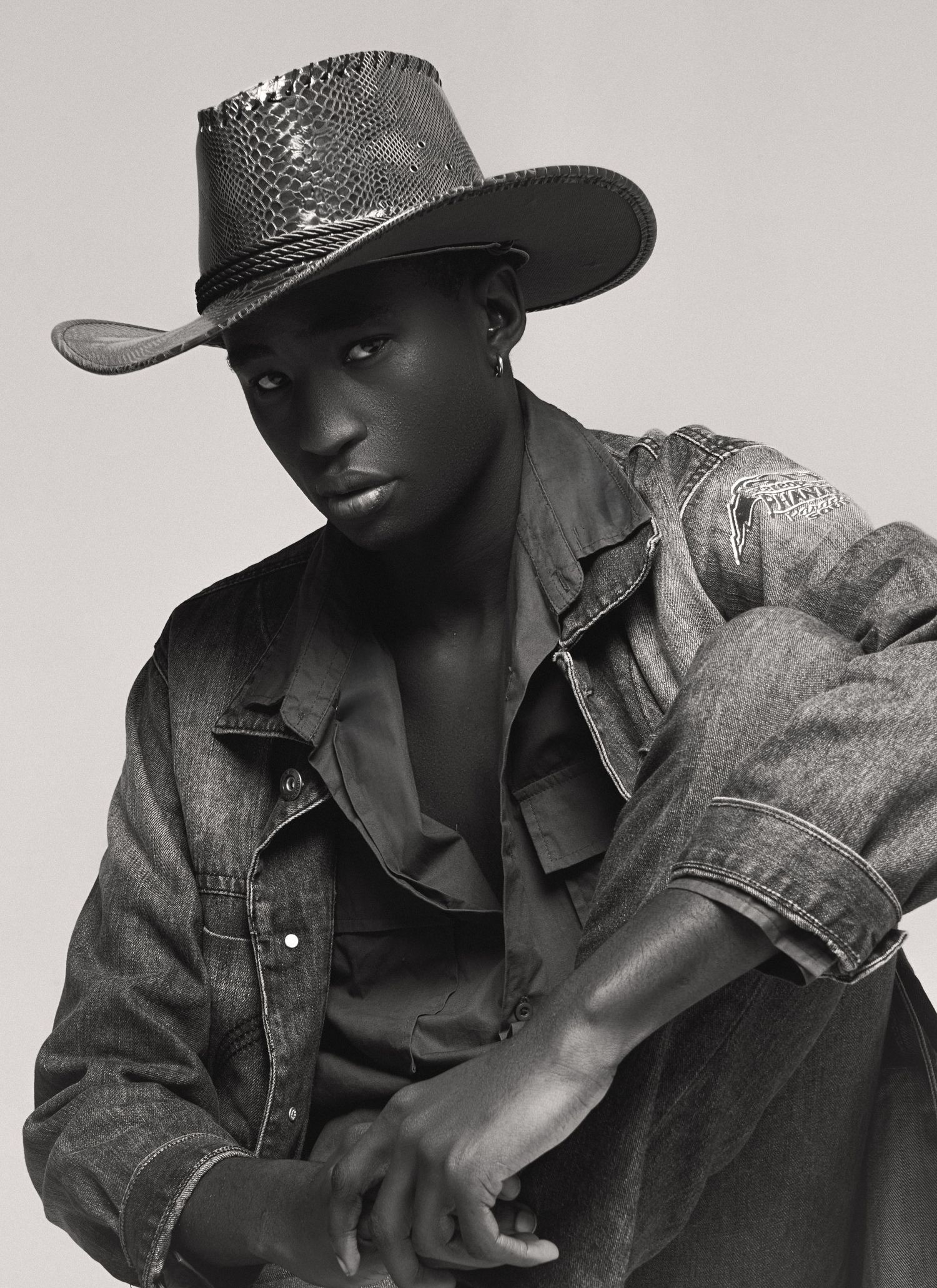 Black-and-white portrait of a male model in a textured cowboy hat and worn denim jacket, posed crouching with arms crossed over his knee.