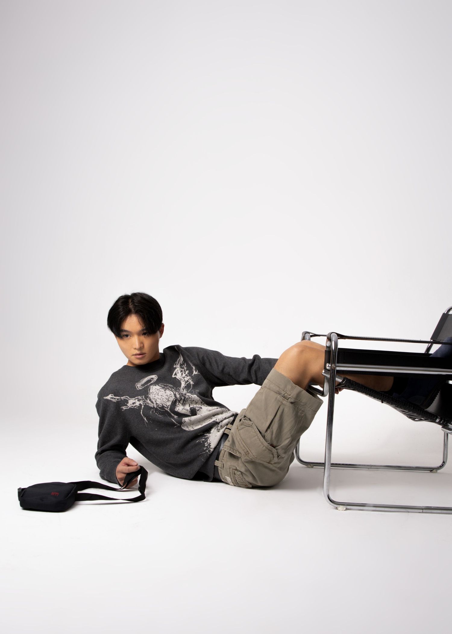 Young man in a graphic sweater and cargo shorts lounges on the studio floor beside a chrome chair and black bag.
