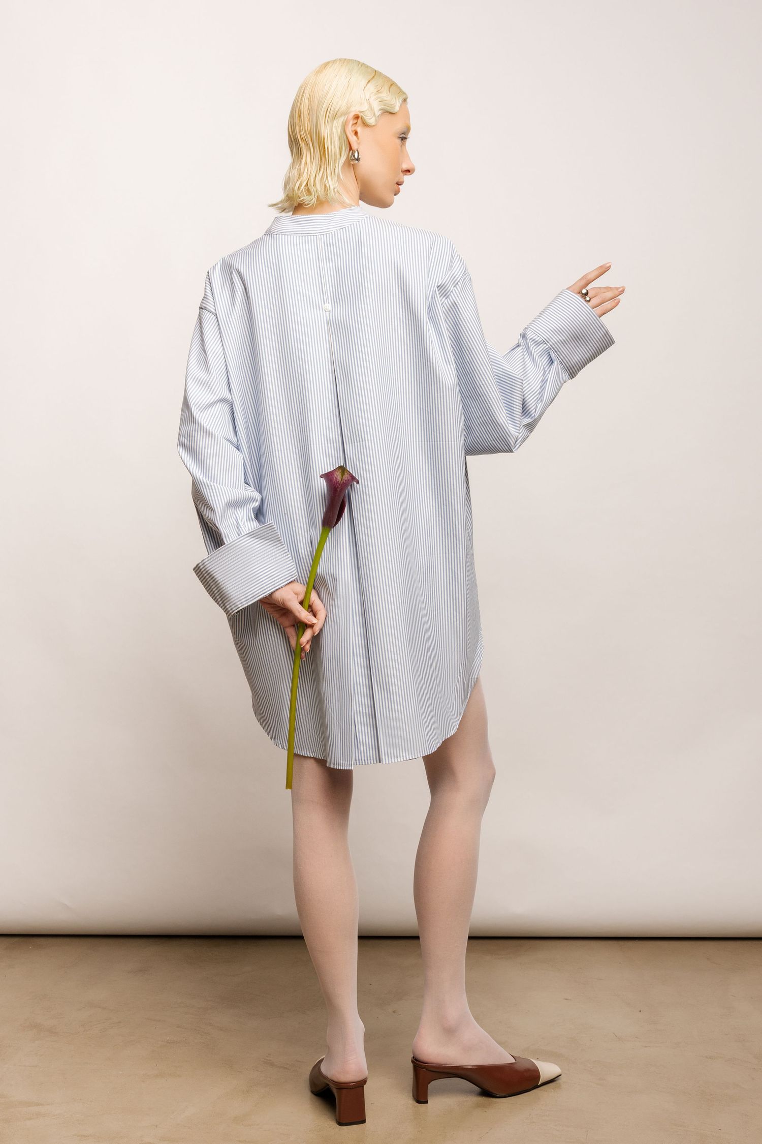 Back view of a model in an oversized blue striped shirt, sheer tights and heels holding a calla lily behind her back.