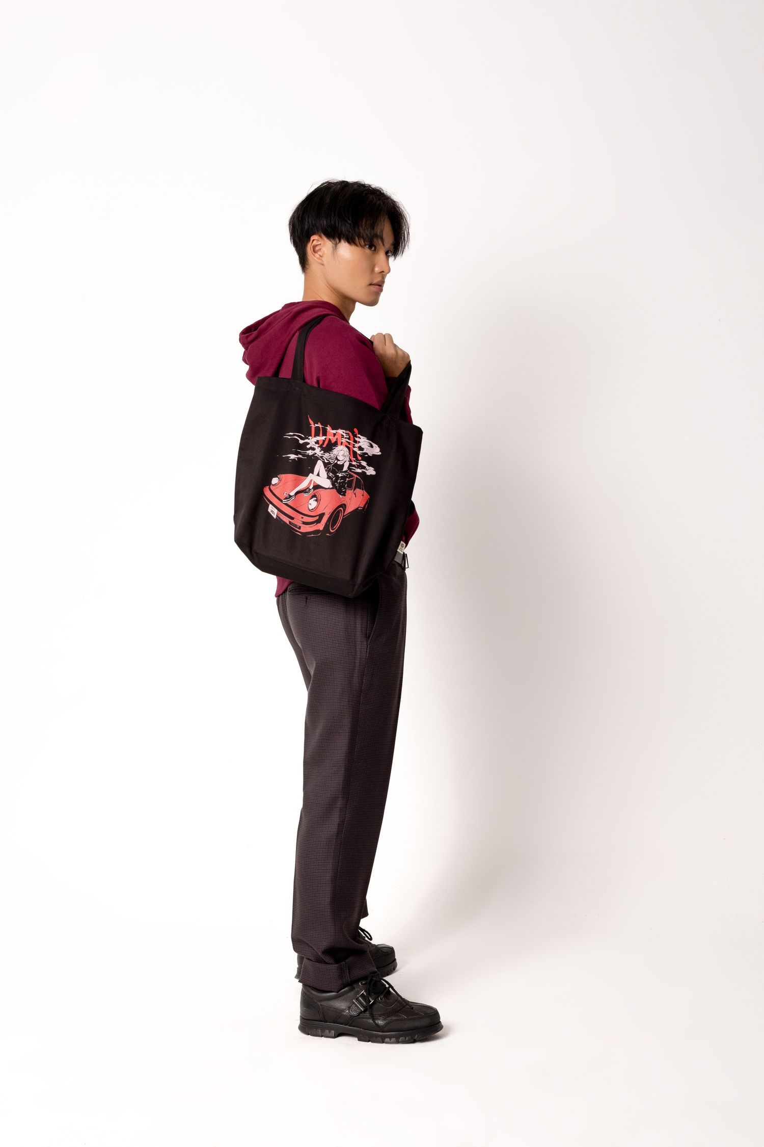Side view of a man in a burgundy hoodie and gray trousers carrying a black tote with a red sports car illustration on his shoulder.