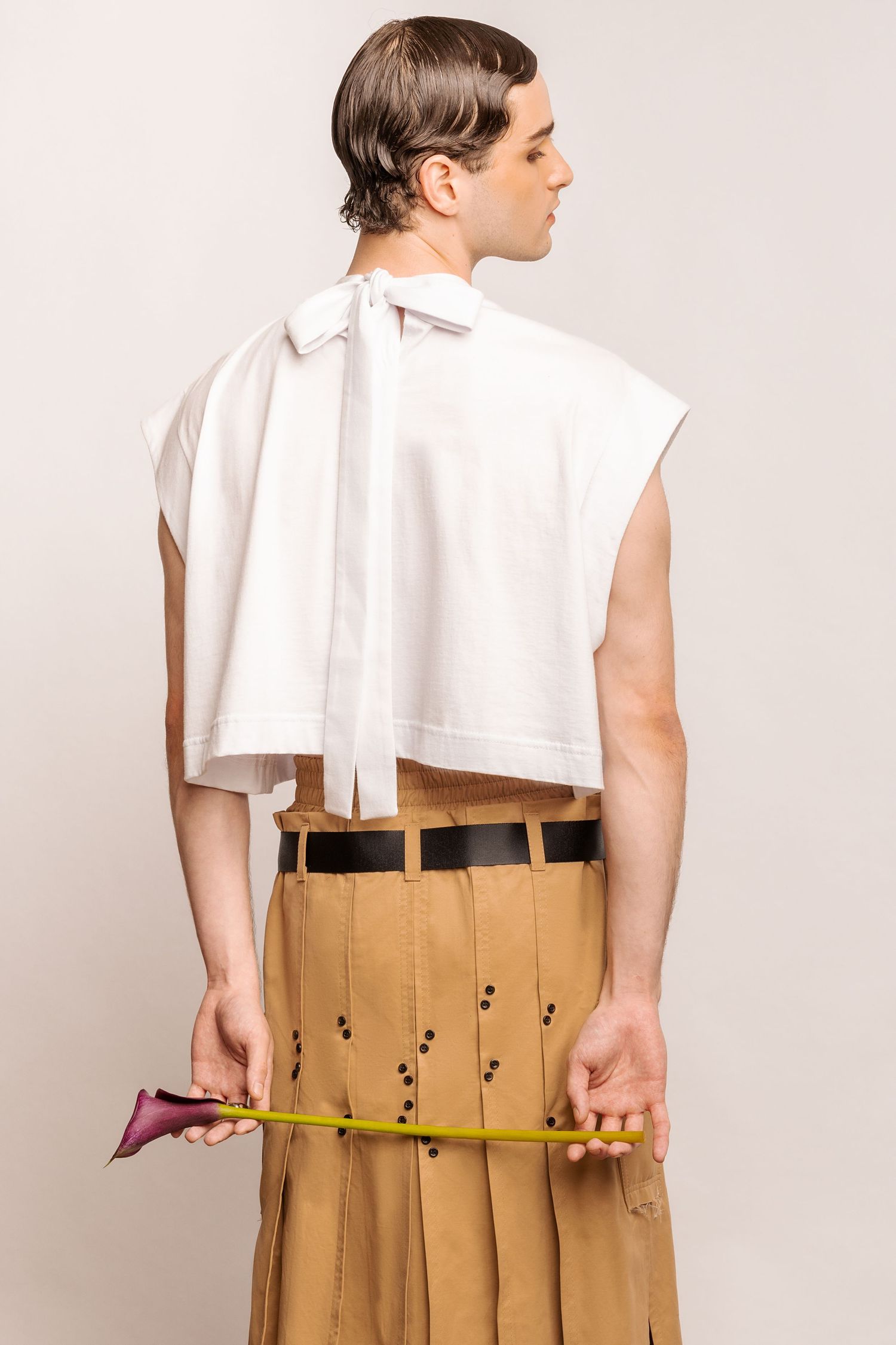 Back view of a model in a white top with a long bow tie, black belt and tan eyelet-detailed pleated kilt, holding a calla lily behind.