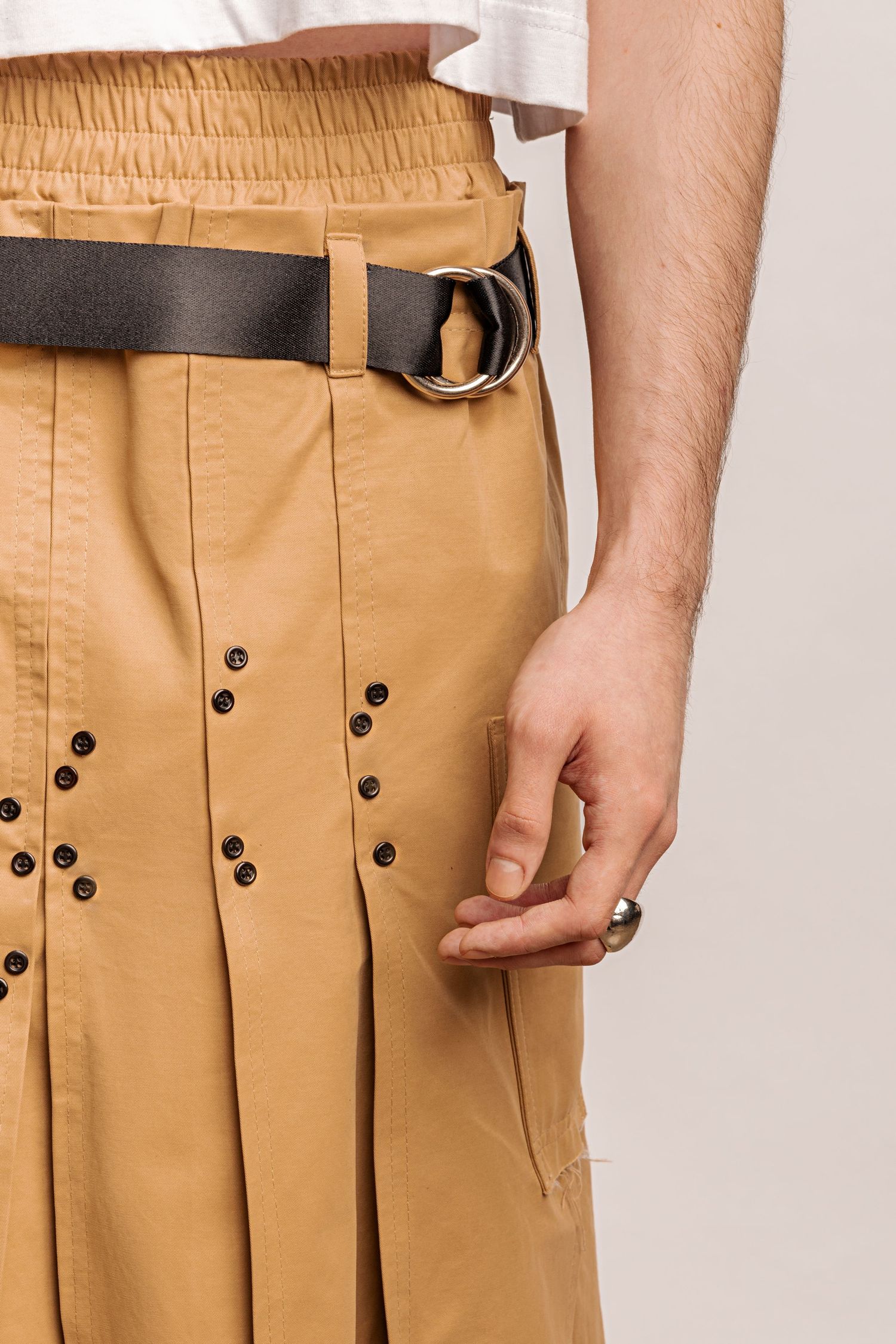 Detail of a tan pleated kilt with metal eyelets, a black belt with a double ring buckle and a hand resting by a pocket.