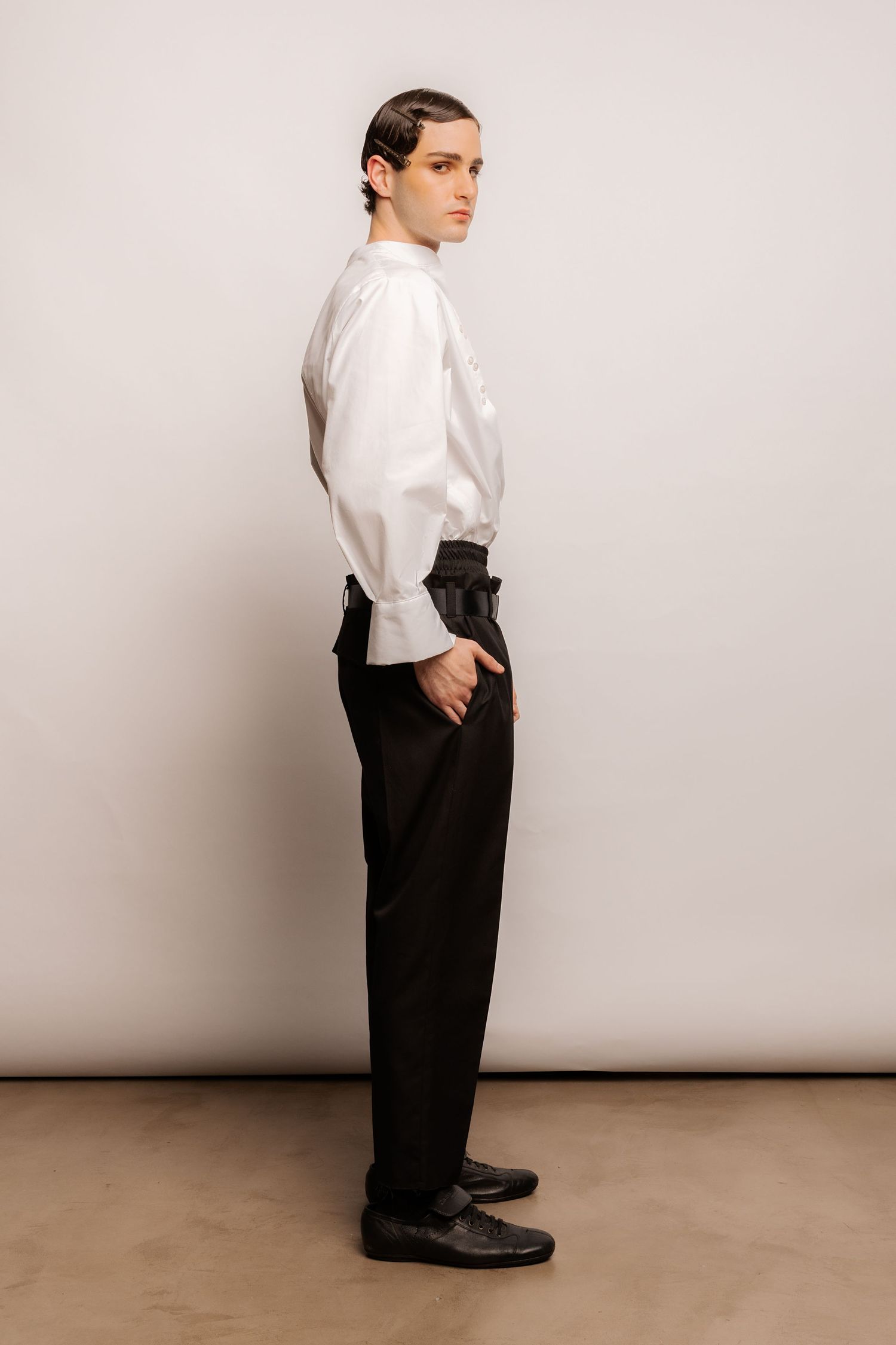 Side profile of a model in a white shirt with gathered sleeves, high-waisted black trousers and black leather shoes, hand in pocket.