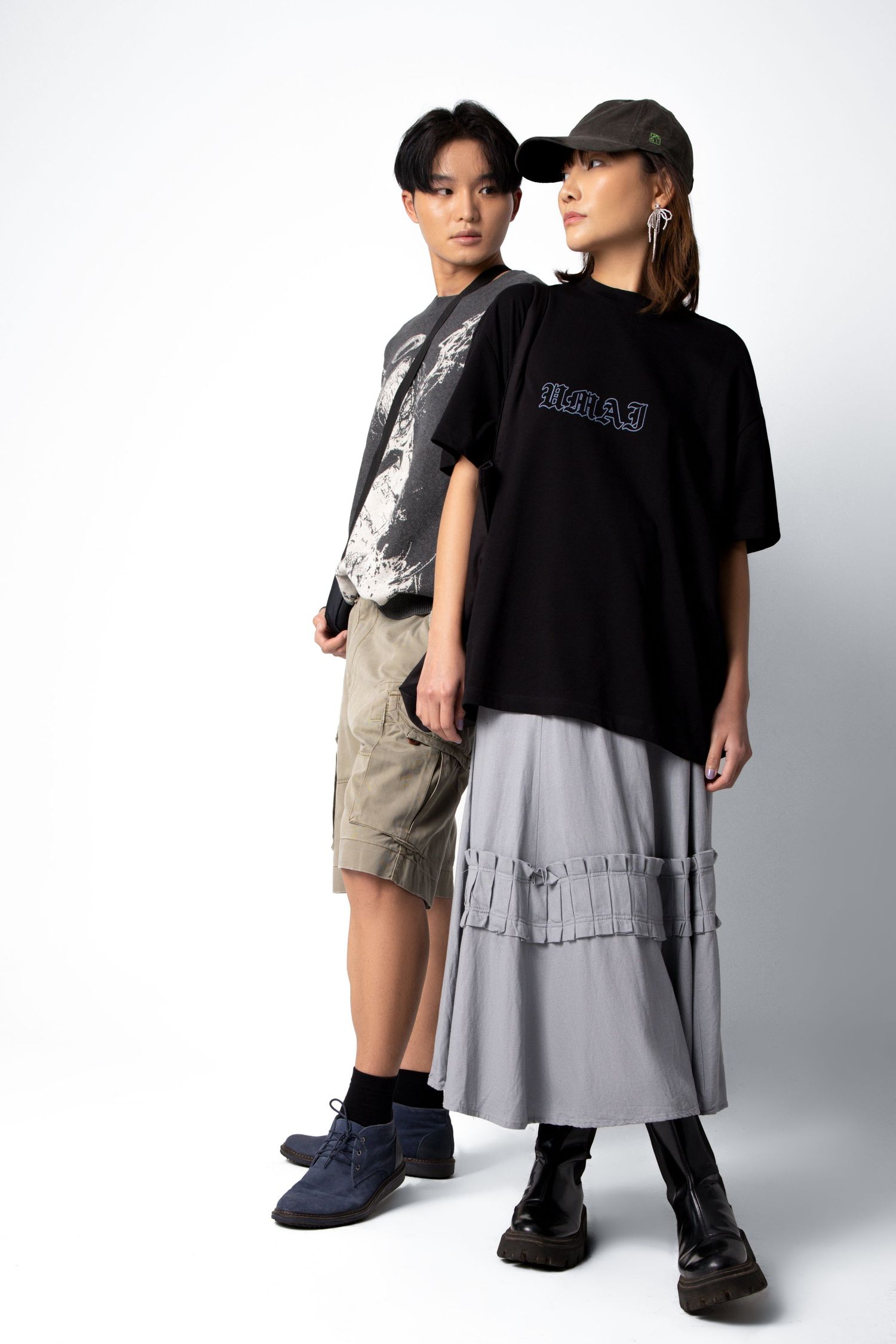 Two models in layered streetwear pose back to back, one in a graphic tee and cargo shorts, the other in a pleated grey skirt.