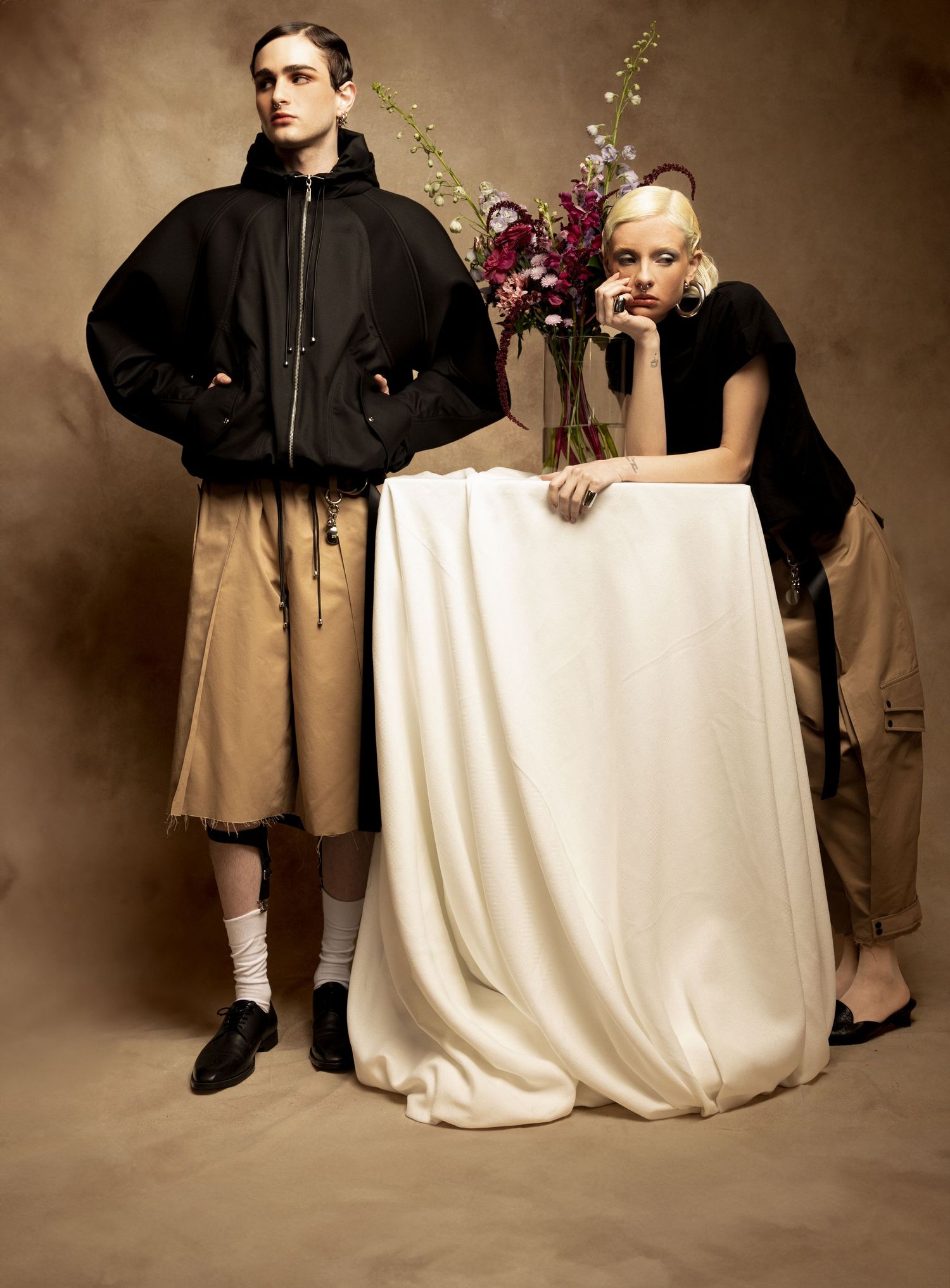 Two fashion models in black hooded jacket, khaki shorts and black tee pose beside a draped white table with a burgundy floral arrangement.