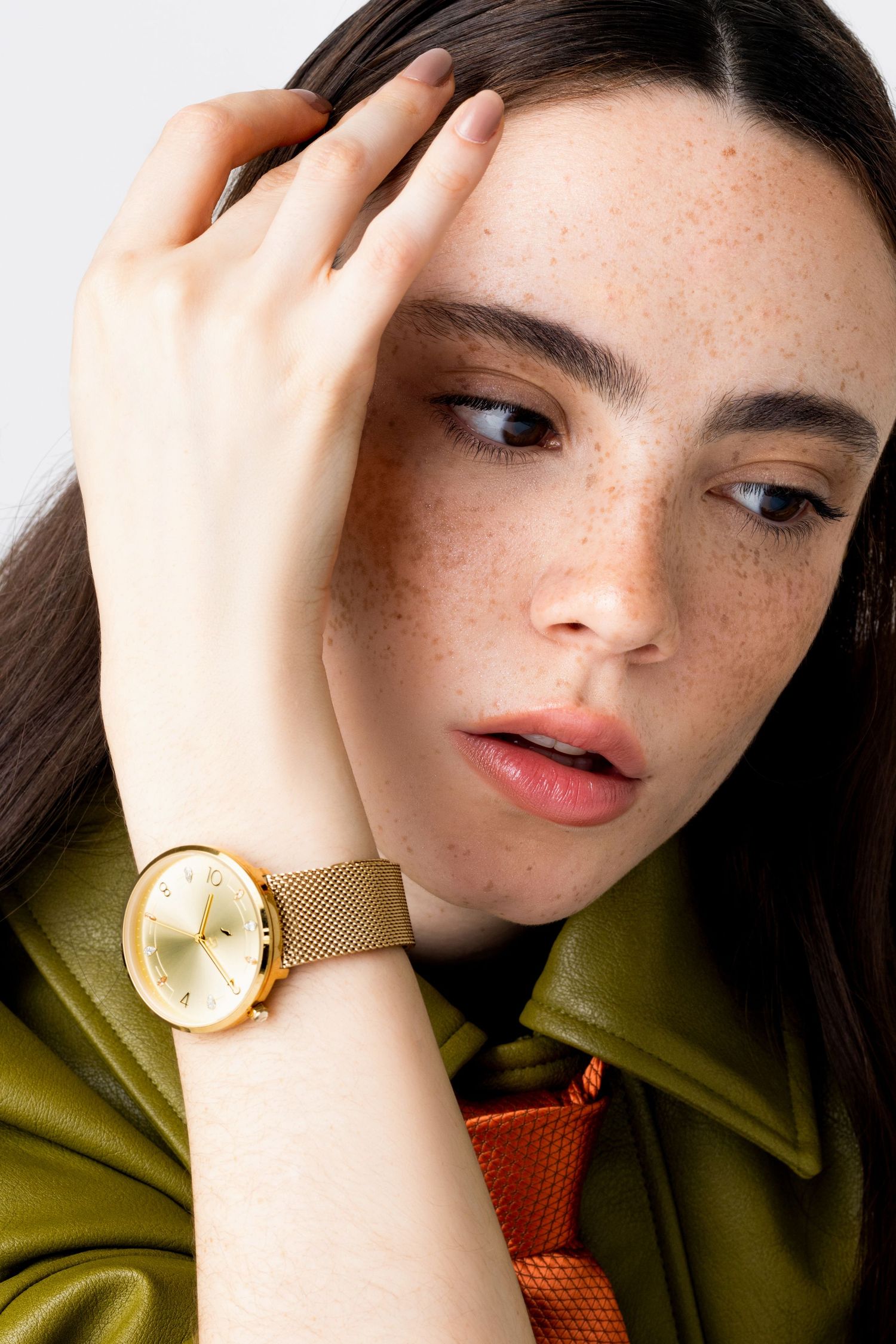 Woman with freckles touches her forehead, wearing a round gold watch with a gold mesh strap and an olive green leather jacket.