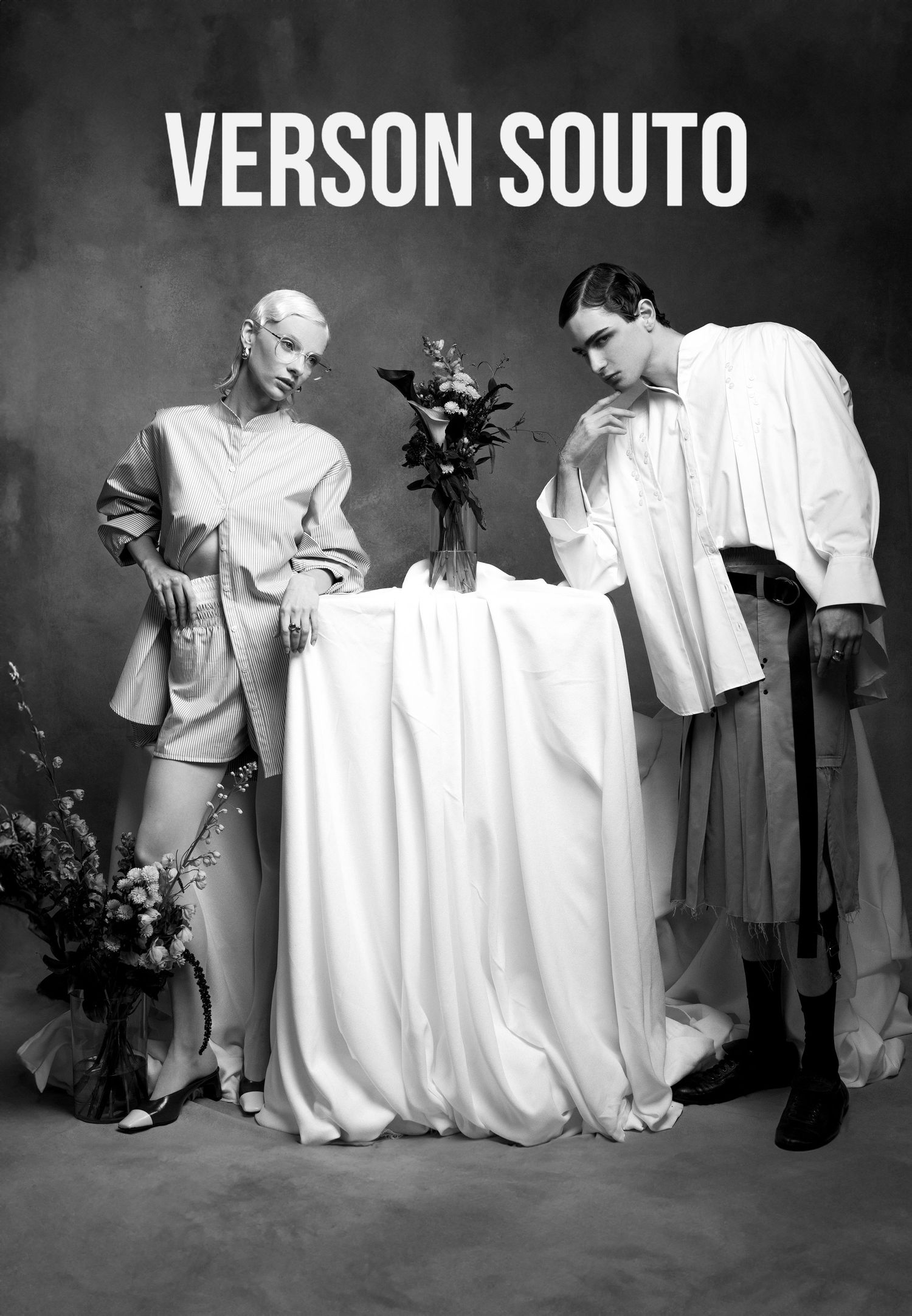Black and white Verson Souto editorial of two models in oversized shirts and a pleated skirt posing around a draped table with flowers.