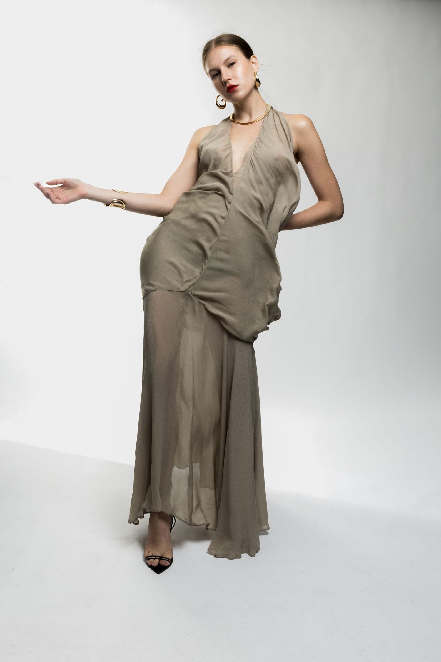Full-length studio photo of a model in an olive sheer halter maxi dress with gold jewelry and black heels against a white backdrop.