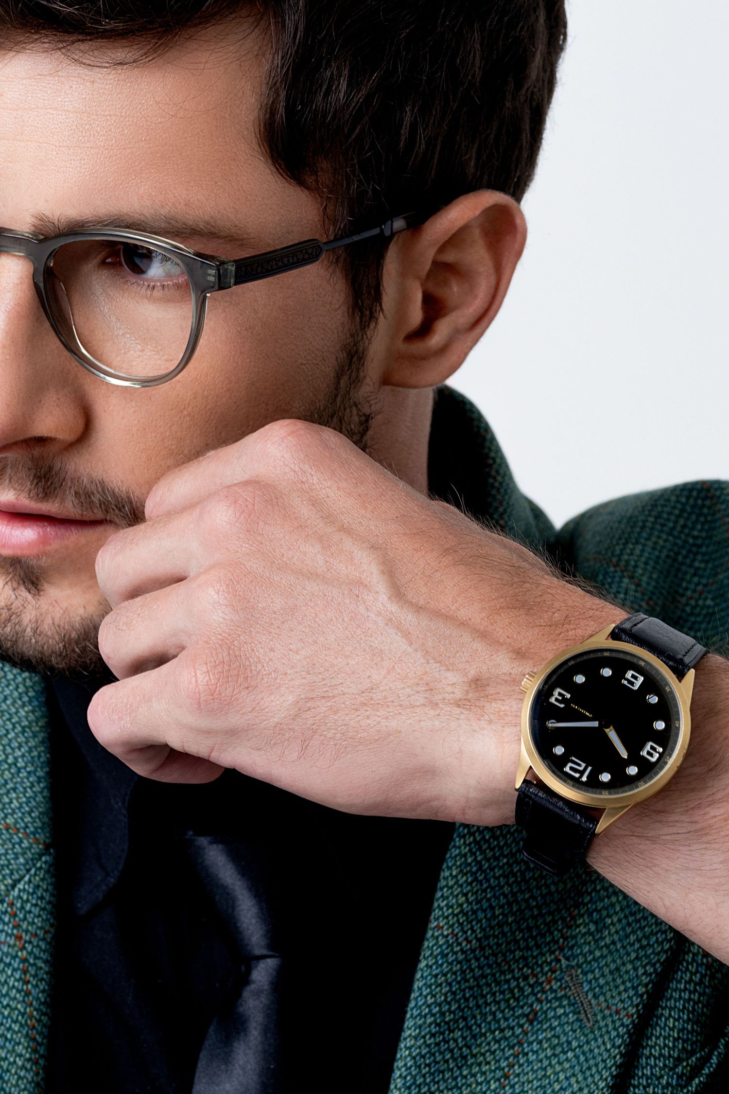 Side profile of a man in glasses and green tweed jacket, hand at his chin, showing a gold watch with a black dial and strap.