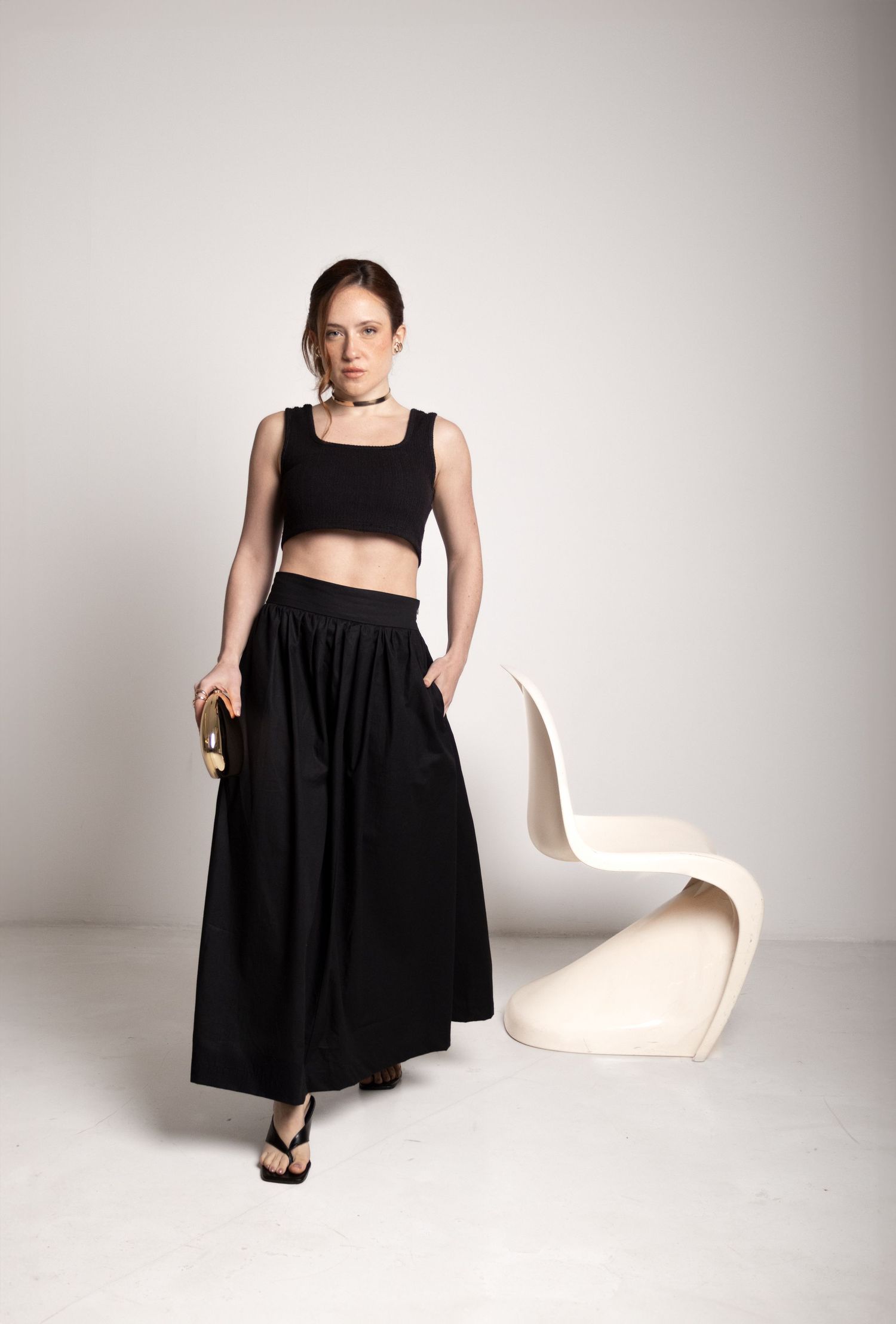 Woman stands in a black crop top and flowing black maxi skirt with hand in pocket beside a sculptural cream chair.