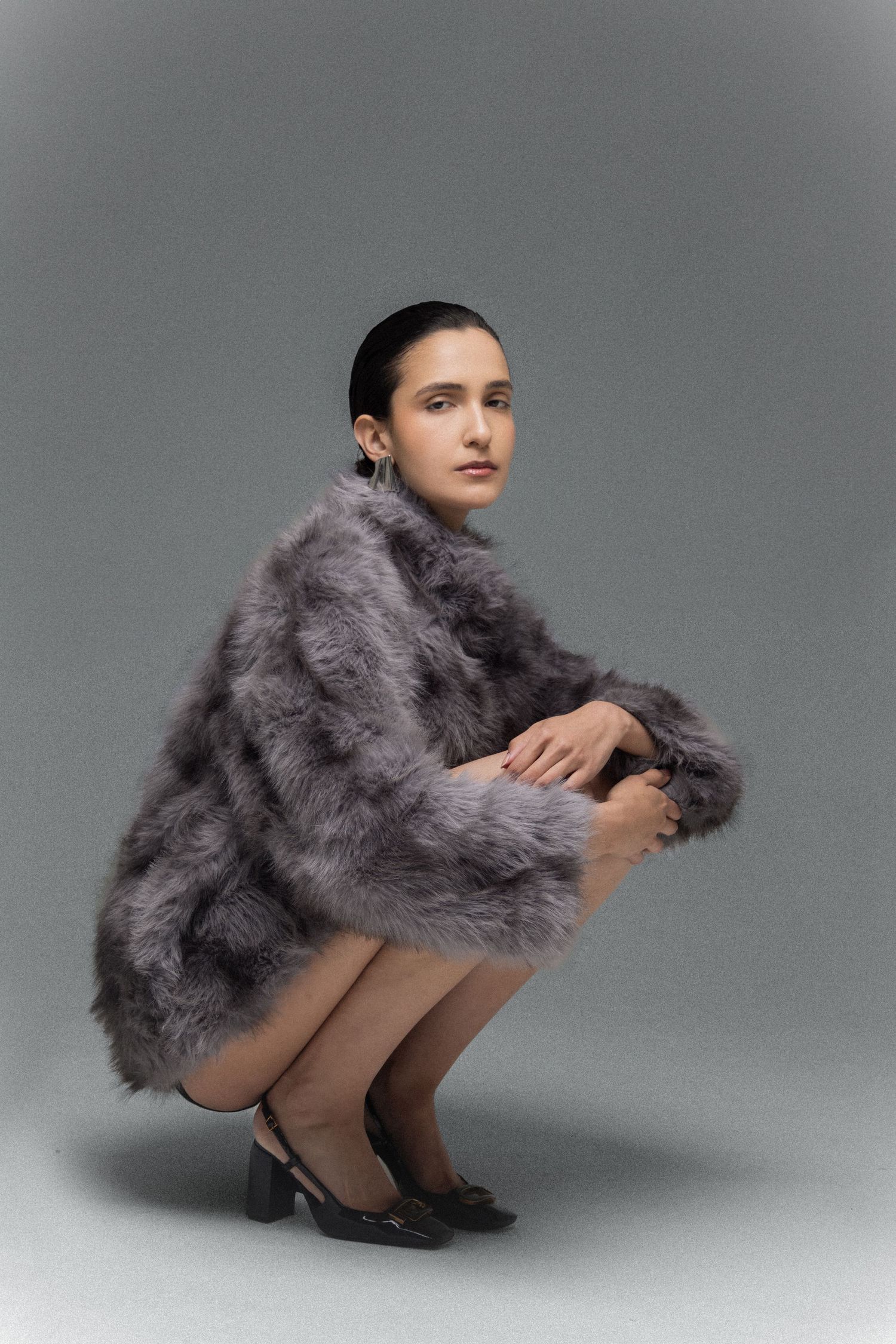 Model crouching in a fluffy gray faux fur coat and black block-heel slingback shoes against a gray studio backdrop.