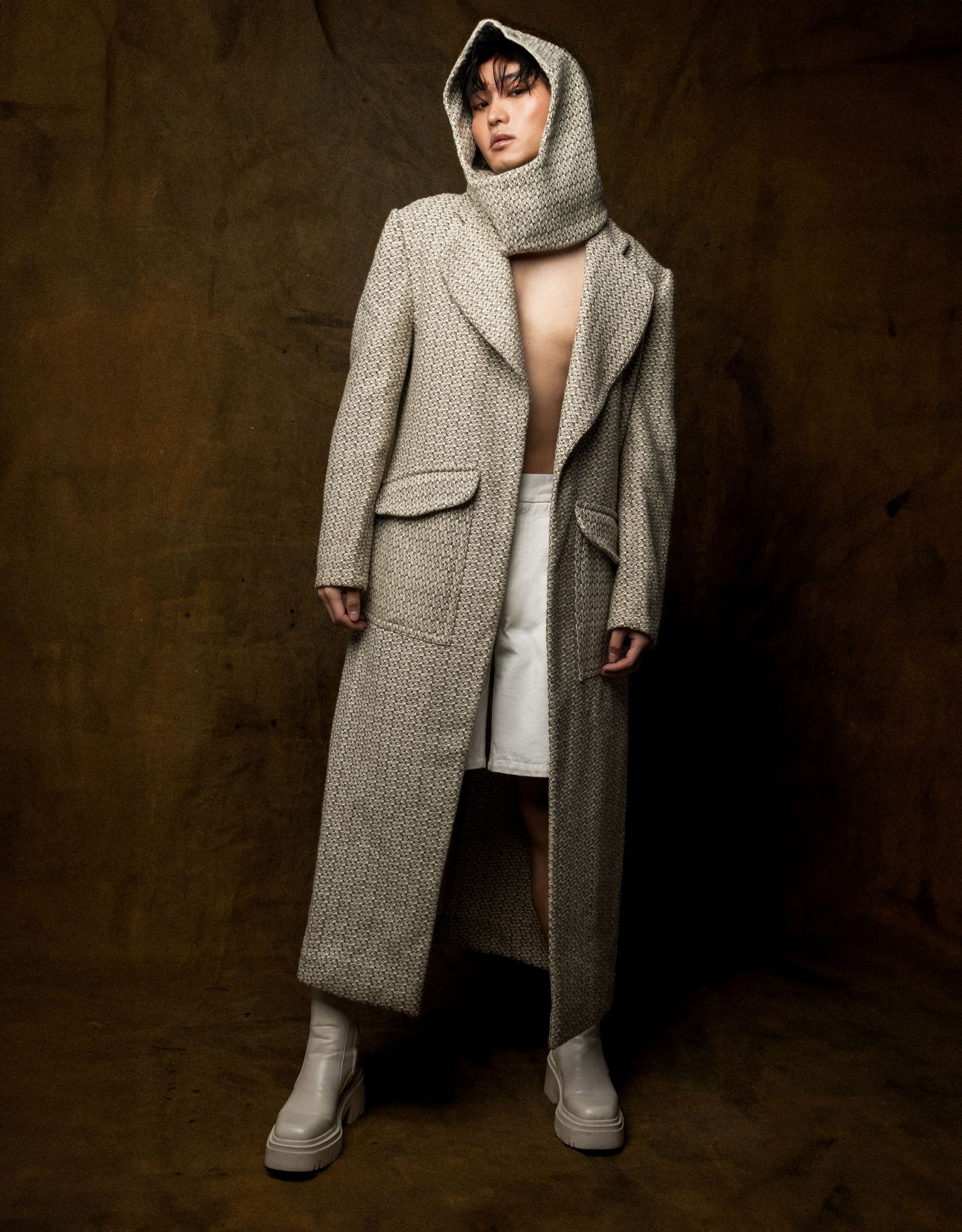 Model stands in an open long tweed coat with attached hood, white skirt and chunky boots, photographed against a dark painterly background.