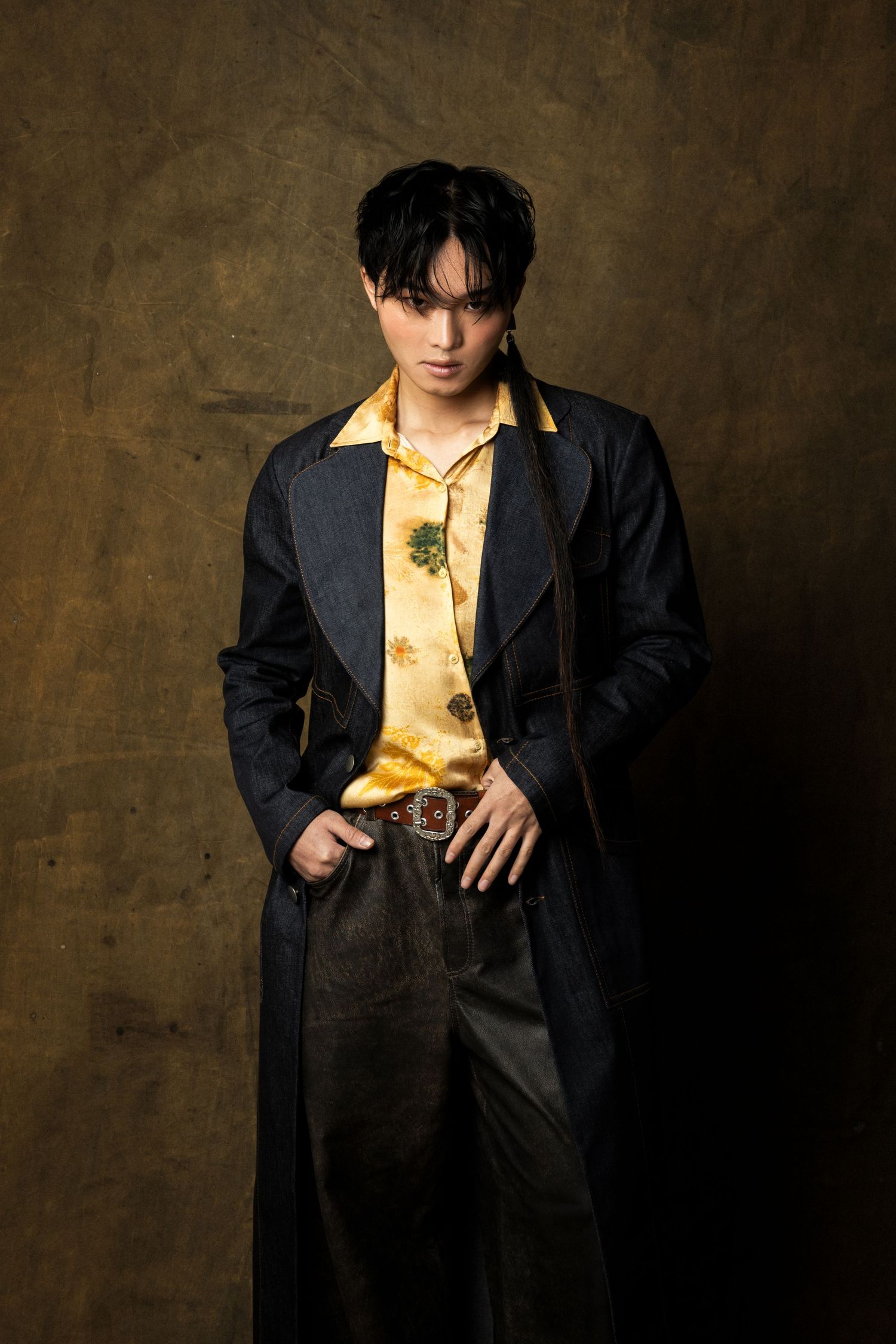 Model with dark hair wears a long denim coat over a yellow floral shirt and belted trousers, posing with hands in pockets.