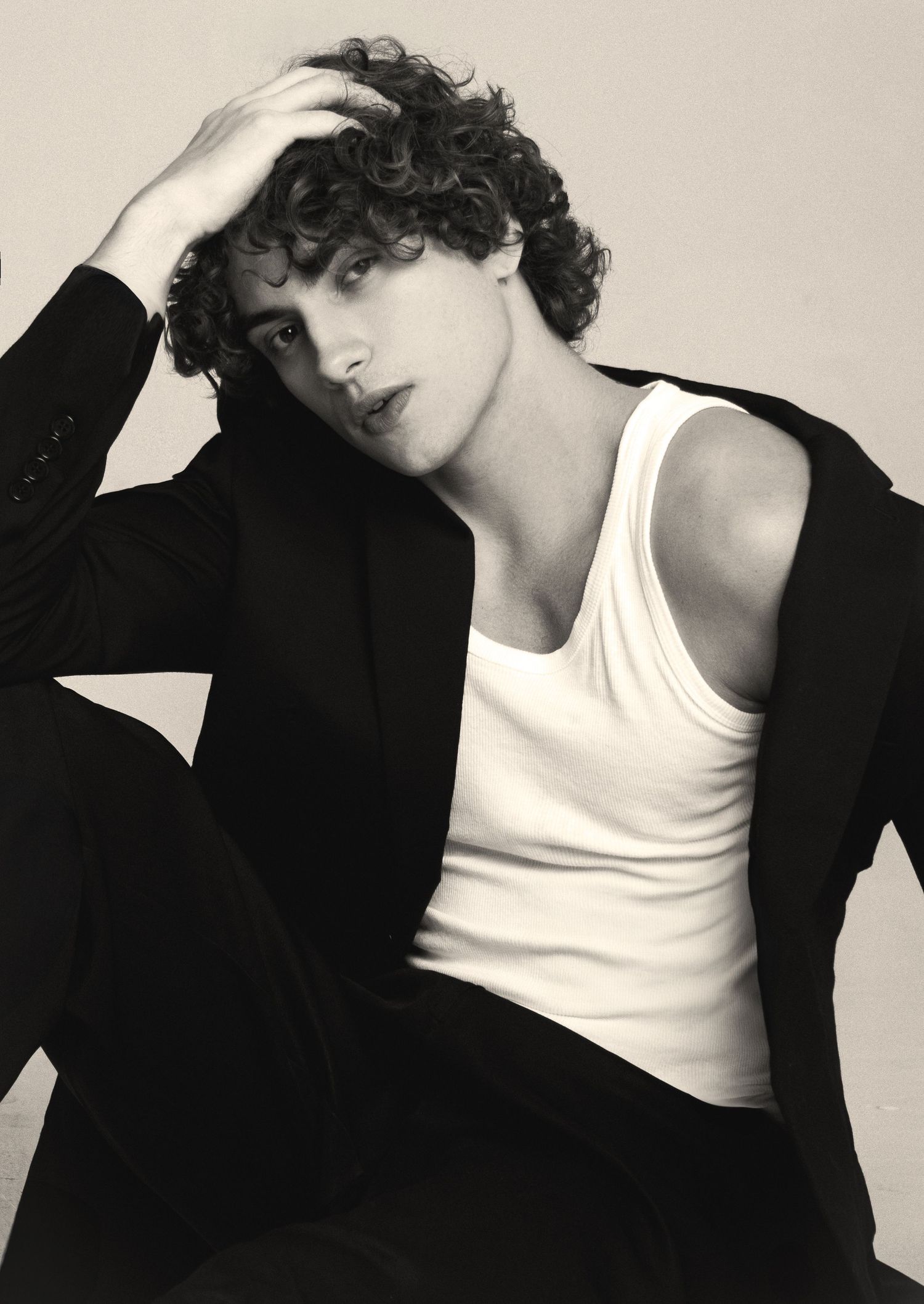 Black and white portrait of a curly-haired young man in a white tank top and black blazer, resting his hand on his head.