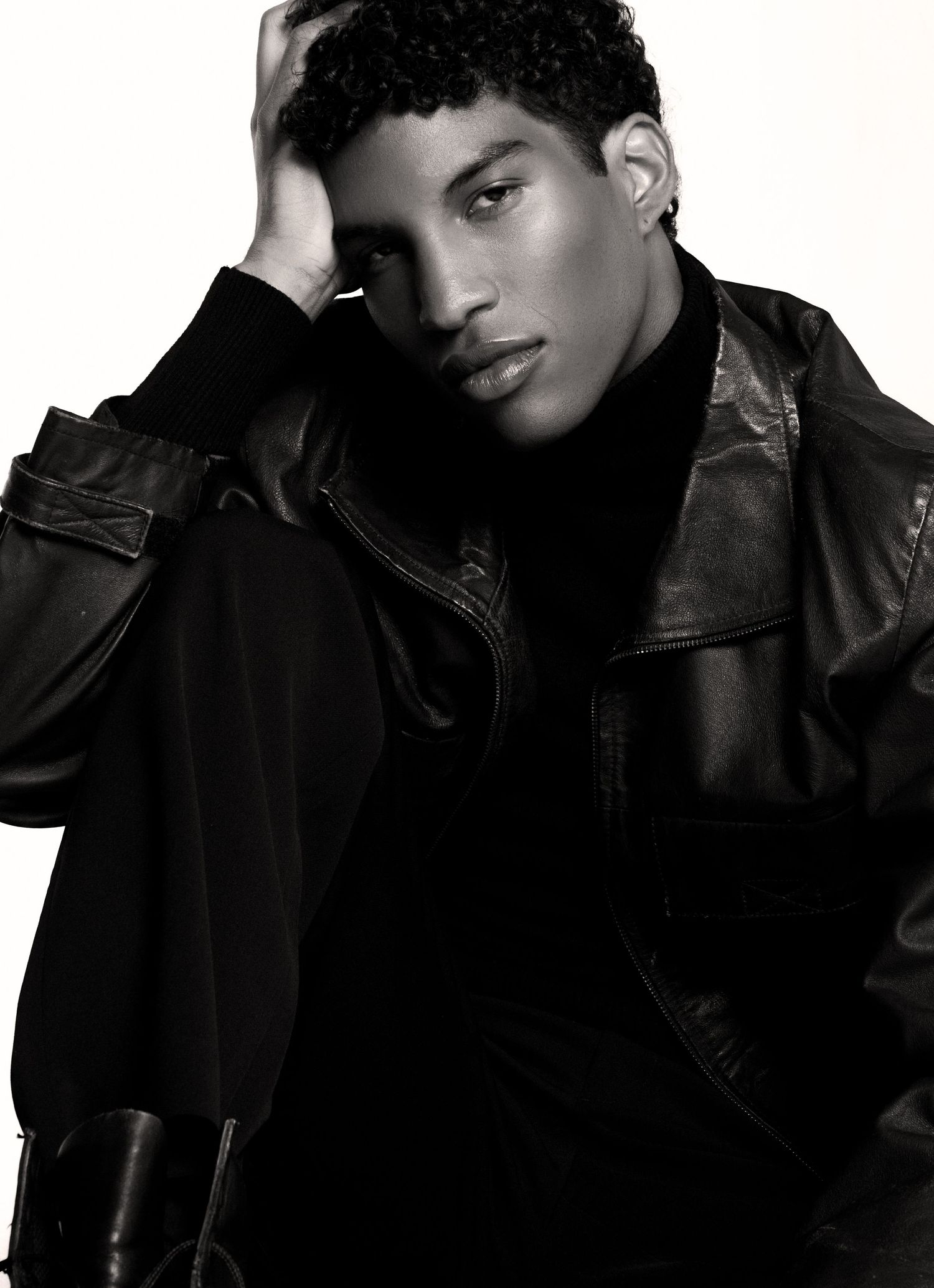 Black and white portrait of a model with curly hair resting his head on his hand, wearing a black leather jacket and turtleneck.