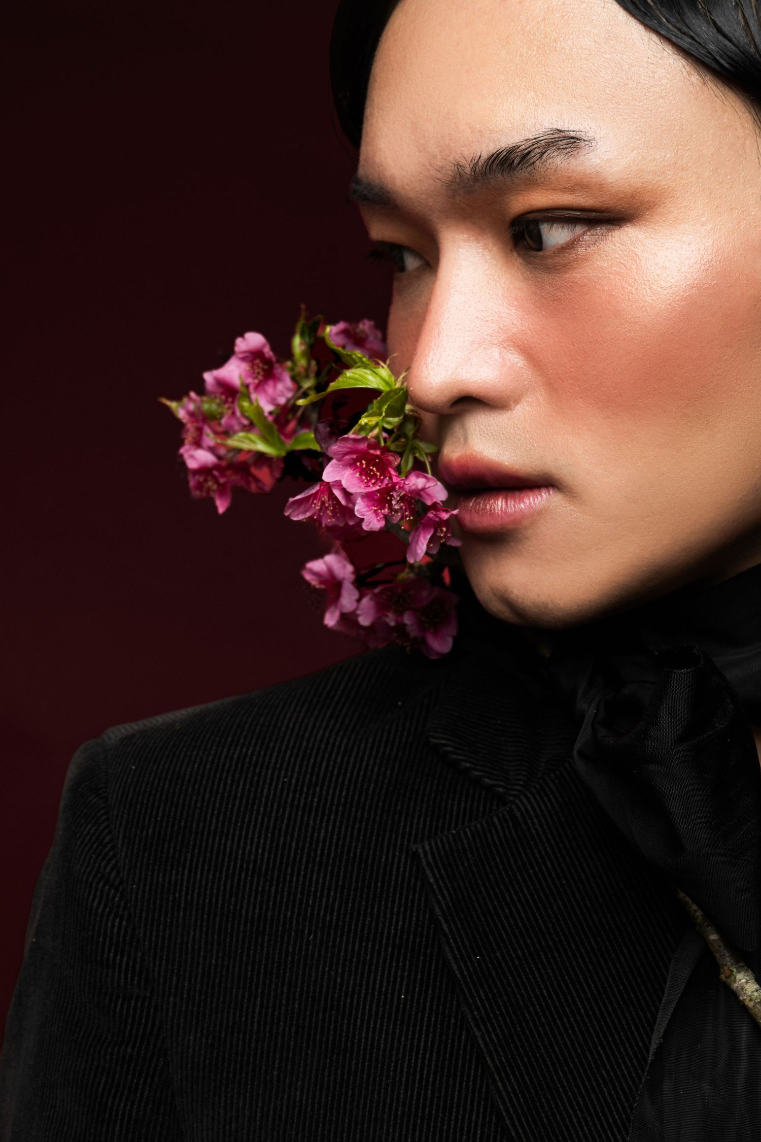 Close-up beauty portrait of a model in profile with glowing makeup, pink blossoms near the cheek, and a black corduroy jacket with ribbon.