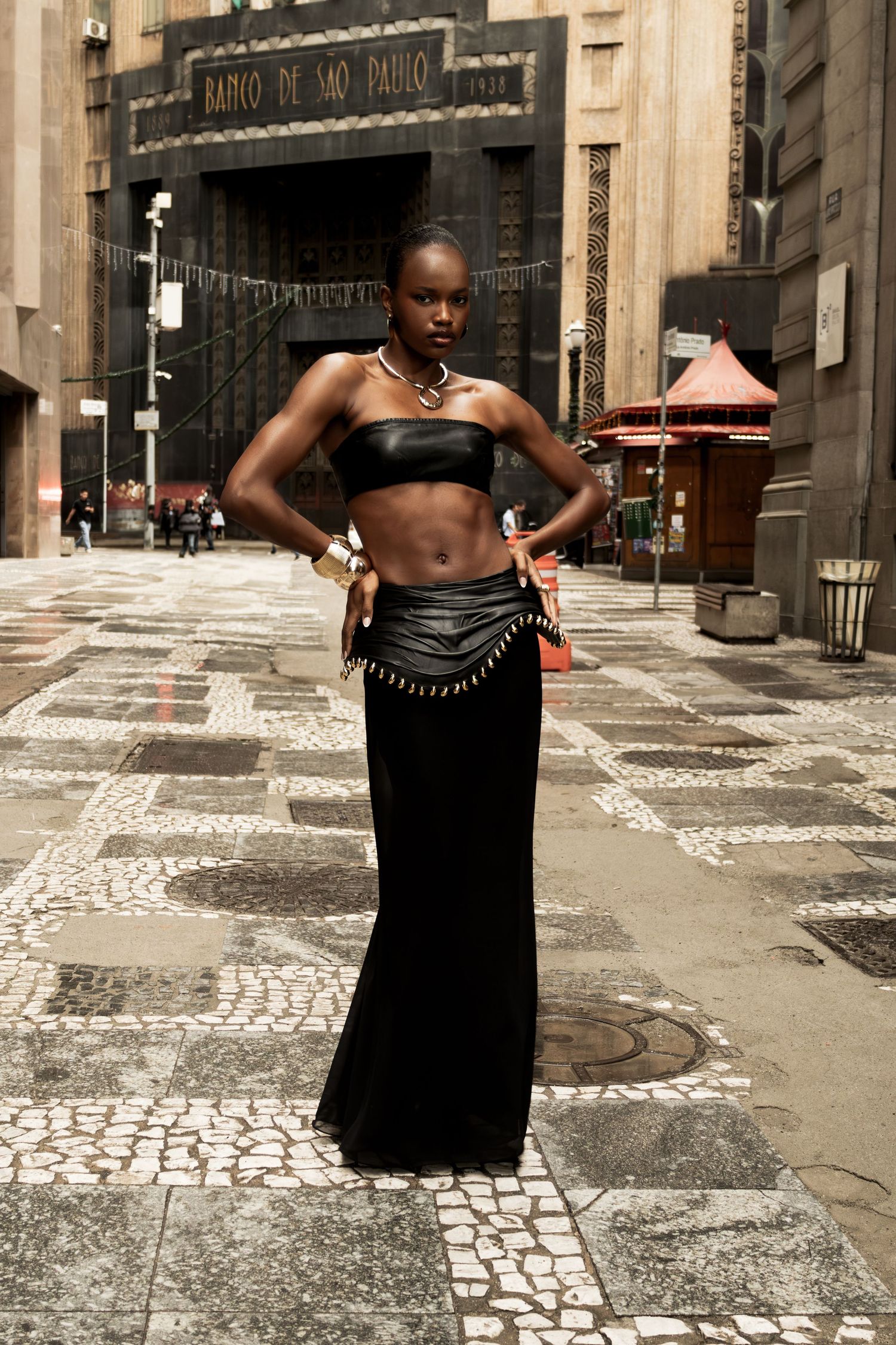Woman in a black leather bandeau top, studded belt and maxi skirt stands on a patterned plaza in front of a historic building.