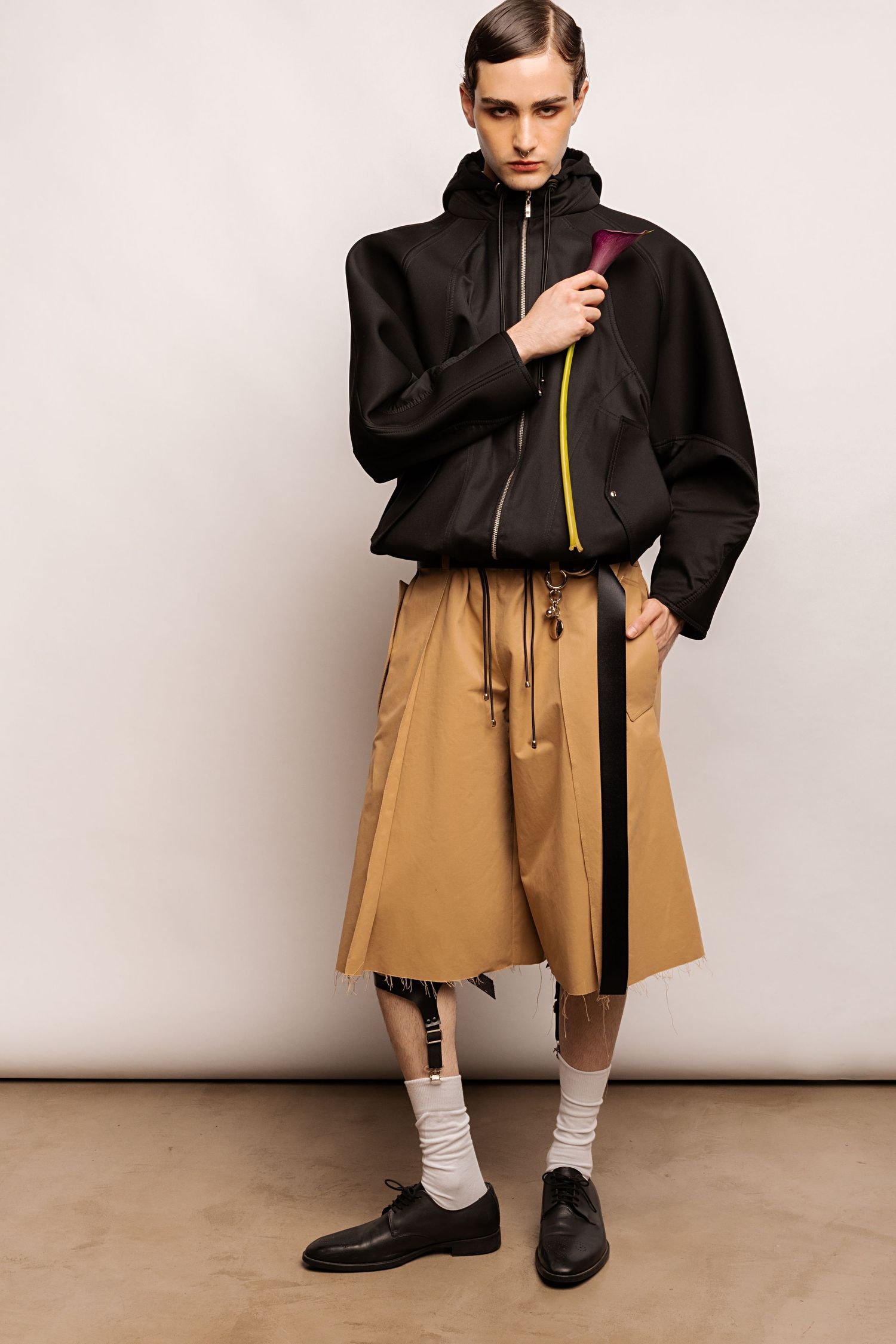 Model poses in a black zip-up hooded jacket with yellow zipper detail, tan wide shorts, white socks and black leather derby shoes.