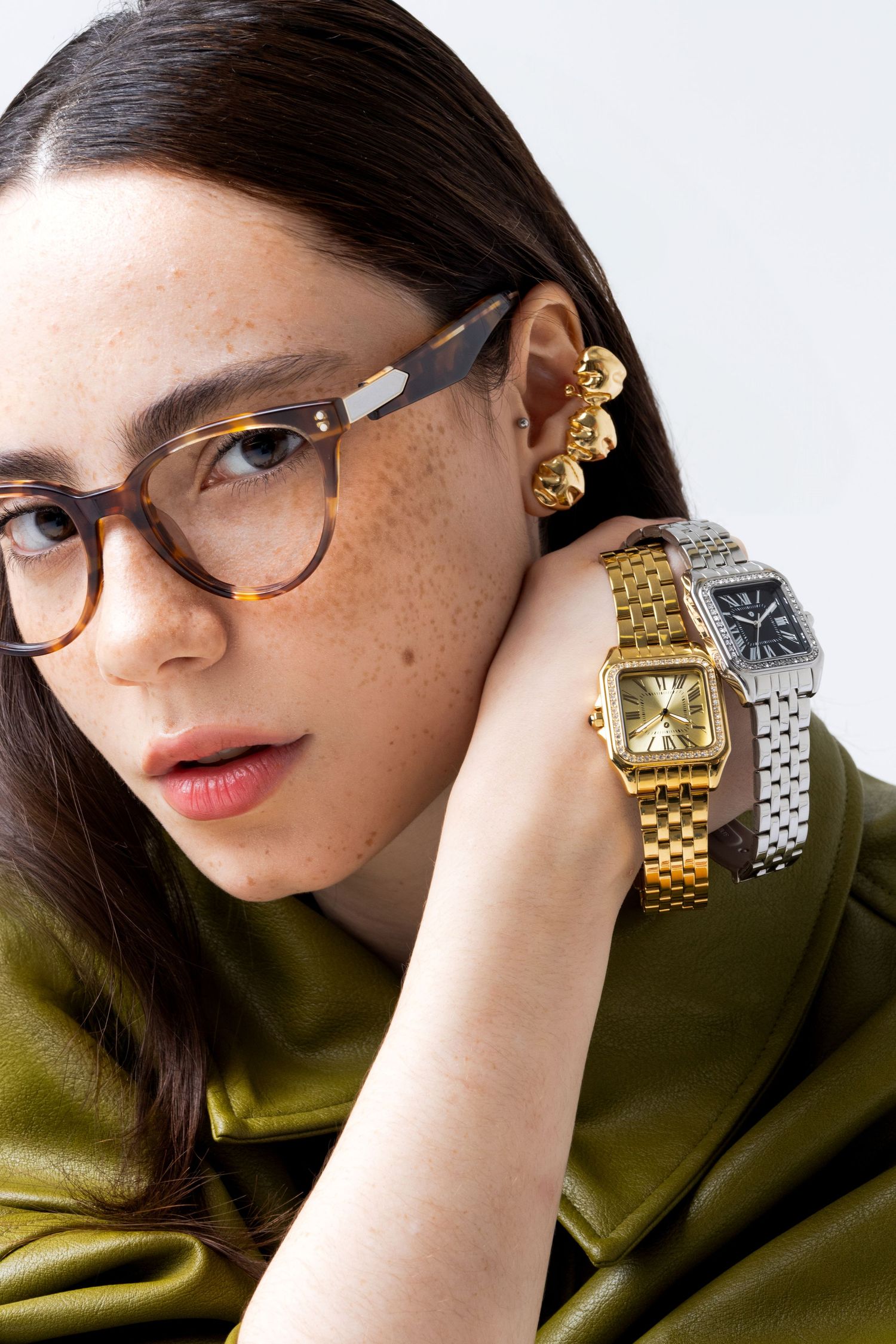 Woman in tortoiseshell glasses stacks a gold rectangular watch and a silver watch with a black dial on her wrist, with a gold ear cuff.