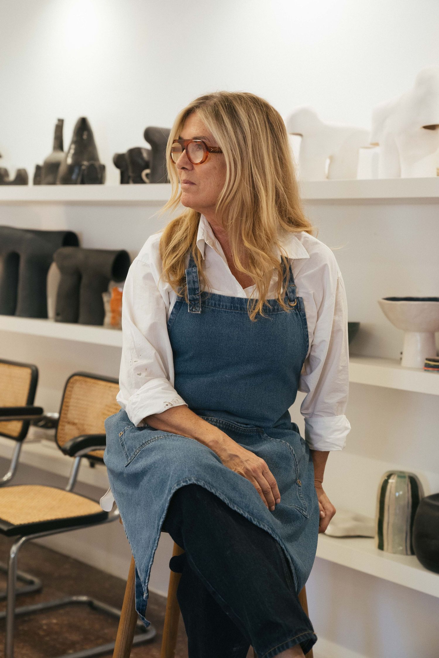 Woman with blonde hair and tortoiseshell glasses in a denim apron sits on a wooden stool in a bright studio with ceramic pottery shelves.
