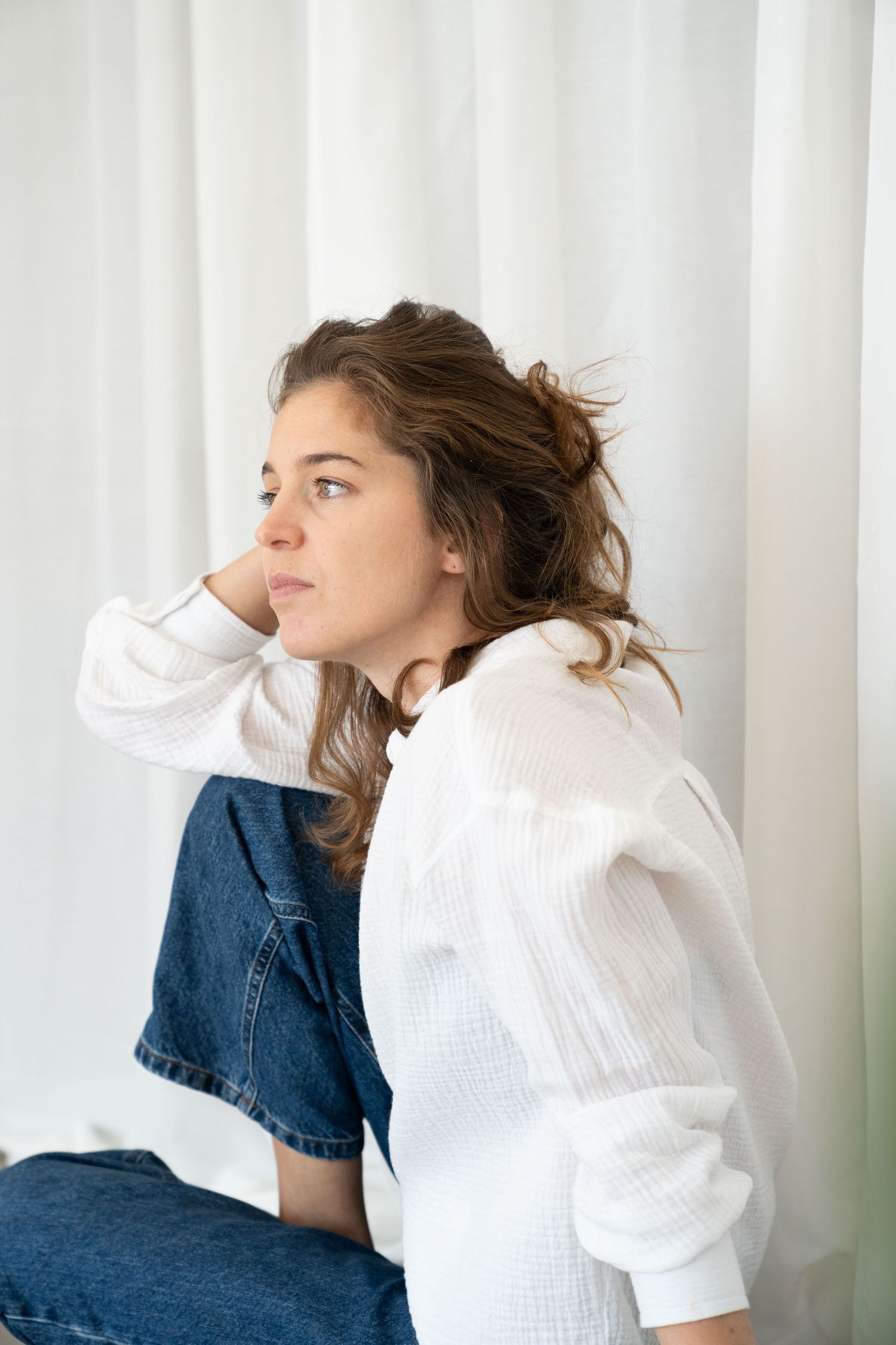 Woman with wavy brown hair, white linen shirt and jeans sits in profile, hand behind her head, before a white curtain.