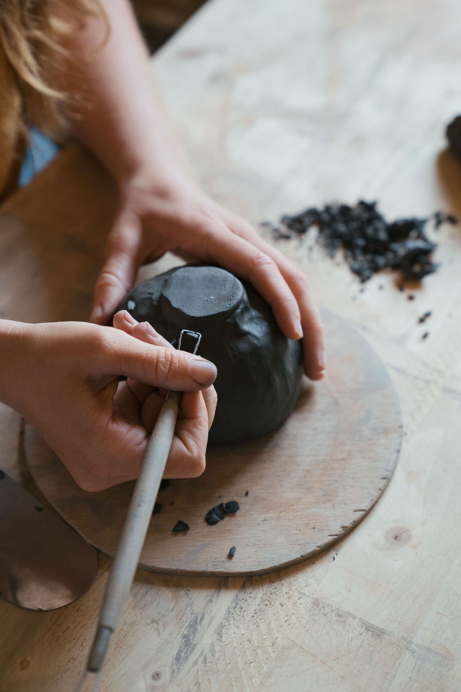 Hands carving a dark clay bowl with a trimming tool on a wooden board, with clay shavings scattered on the table.