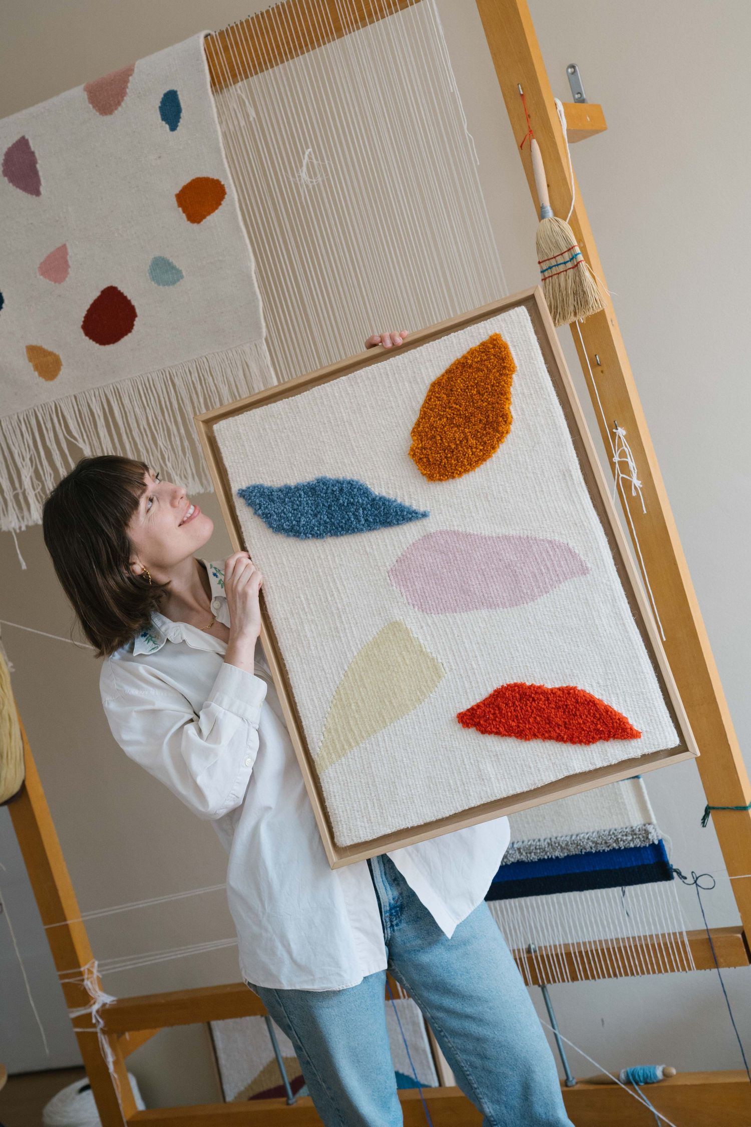 Smiling weaver holds a framed handwoven tapestry with colorful abstract tufted shapes in front of a wooden loom in a bright studio.