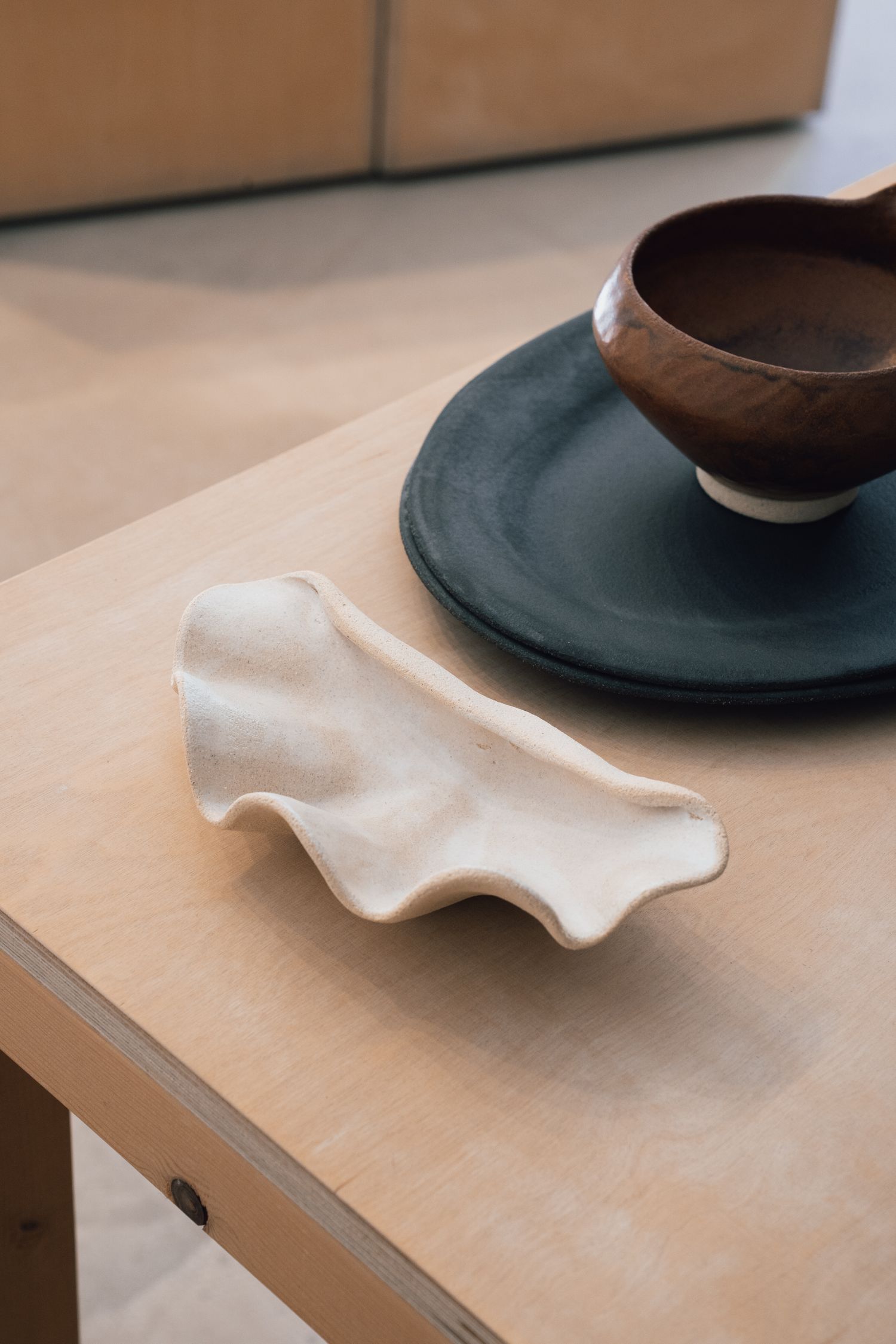 Handmade cream ceramic dish with a wavy edge on a light wooden table beside a dark plate and a brown ceramic bowl.