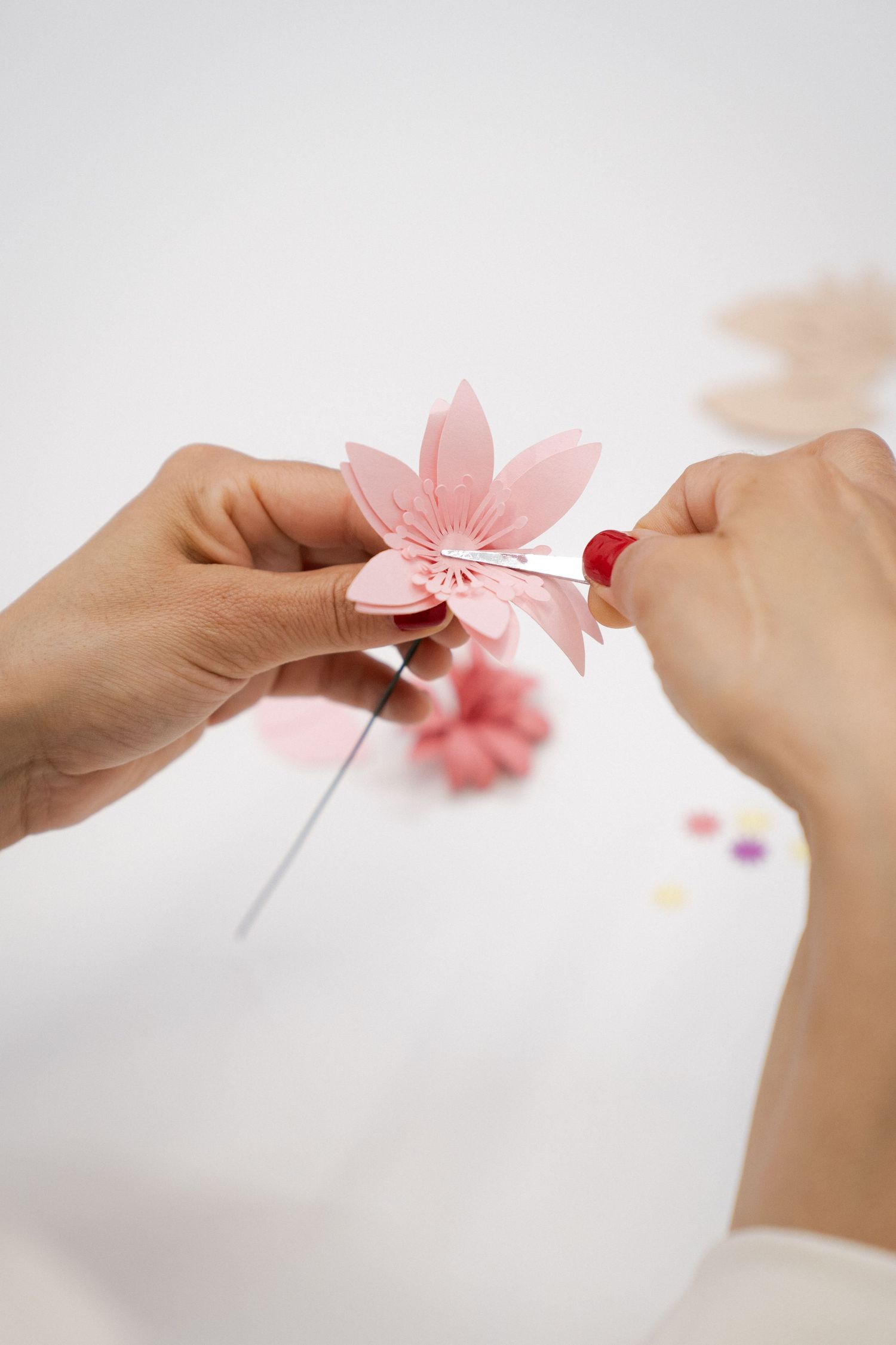 Hands with red nail polish crafting a delicate pink paper flower, with a wire stem and more handmade blossoms on a white table.